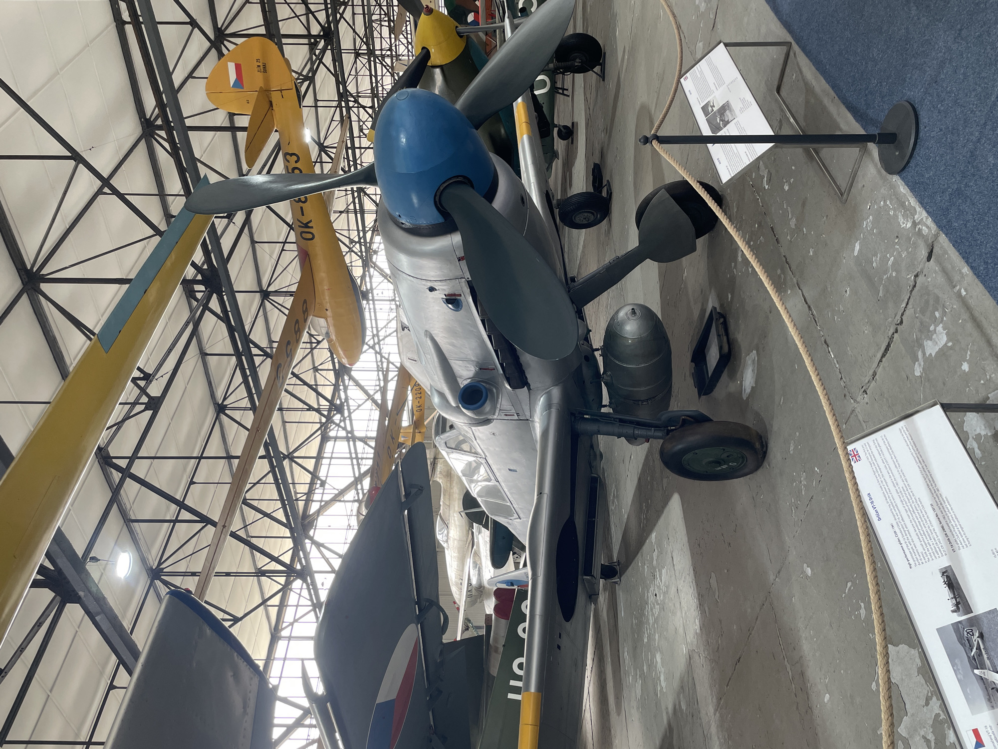 An indoor photograph shows a row of vintage aircraft parked inside a hangar. In the foreground, a silver-colored propeller aircraft with a blue nose cone is prominently displayed. It has a single engine and a two-bladed propeller. Visible under the main aircraft is a black drip tray. Further back, other aircraft are visible, including a yellow-colored plane with the registration "OK-853" on its wing and tail, and another with a green and yellow striped wing. The hangar has a white ceiling supported by a dark metal truss structure. A rope barrier separates the viewer from the aircraft, and to the right, a display board with text and images about the aircraft is positioned. The floor is concrete, with some painted lines and stains. The overall lighting suggests an overcast day or artificial illumination within the hangar. The location is identified as the Aviation Museum Kbely in Prague, Czech Republic.
