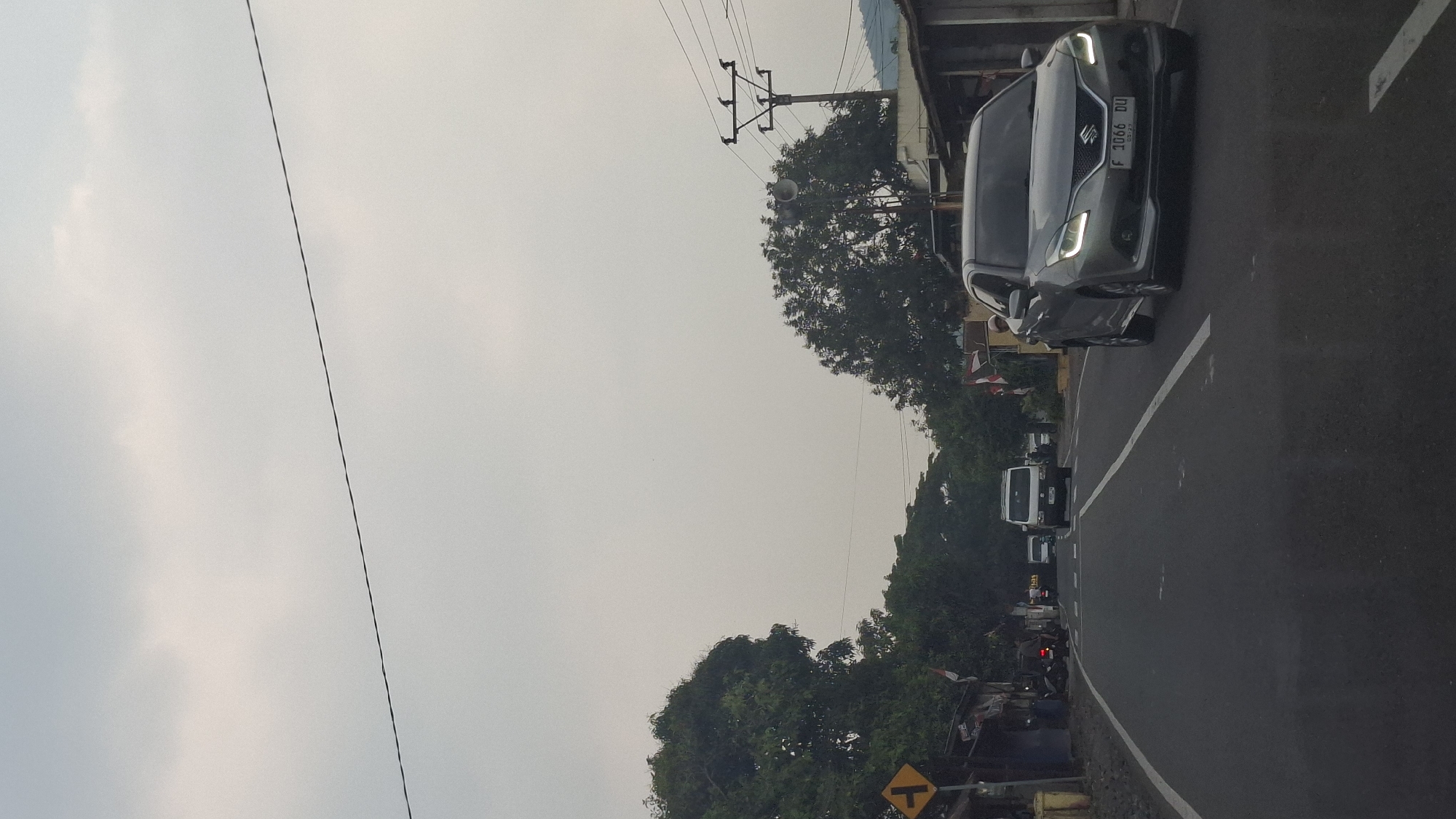 A Suzuki Baleno car, license plate F 1066 DU, travels down a paved road in Susukan, Indonesia. The car is silver and has its headlights on. A white van is in front of the car. The road is lined with trees and buildings. A traffic sign with a left-turn arrow is visible on the left side of the road. The sky is overcast. There are power lines and a utility pole visible.