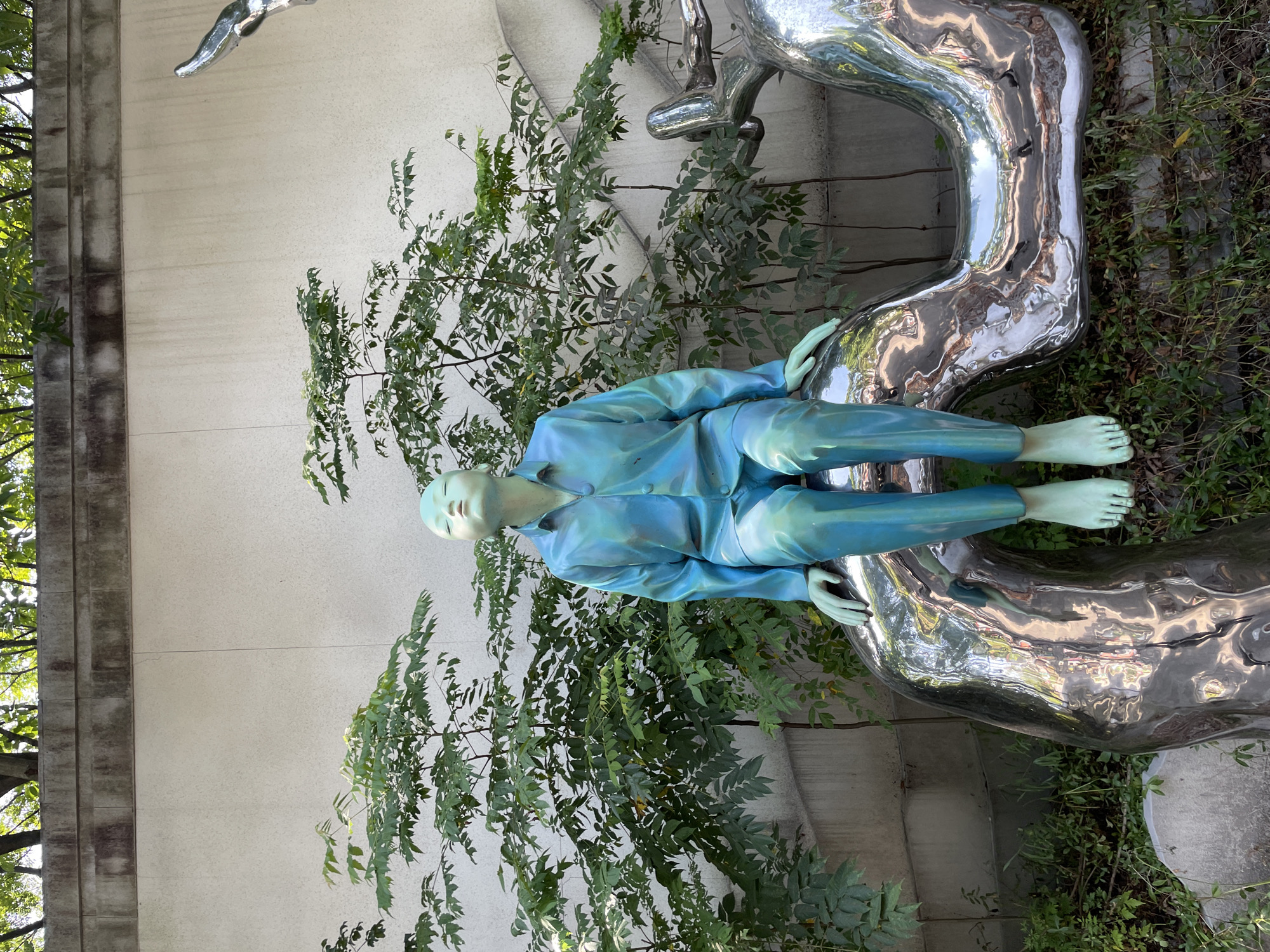 In a courtyard, a turquoise sculpture of a person sits on a chrome structure. The sculpture is wearing a blue-green shirt and pants, with its eyes closed and head tilted back. Green leaves from trees partially obscure the background, which consists of a pale wall and a concrete structure. The chrome sculpture reflects the surroundings, showing distorted images of trees and the sky. The scene is outdoors, with the lighting suggesting daytime.