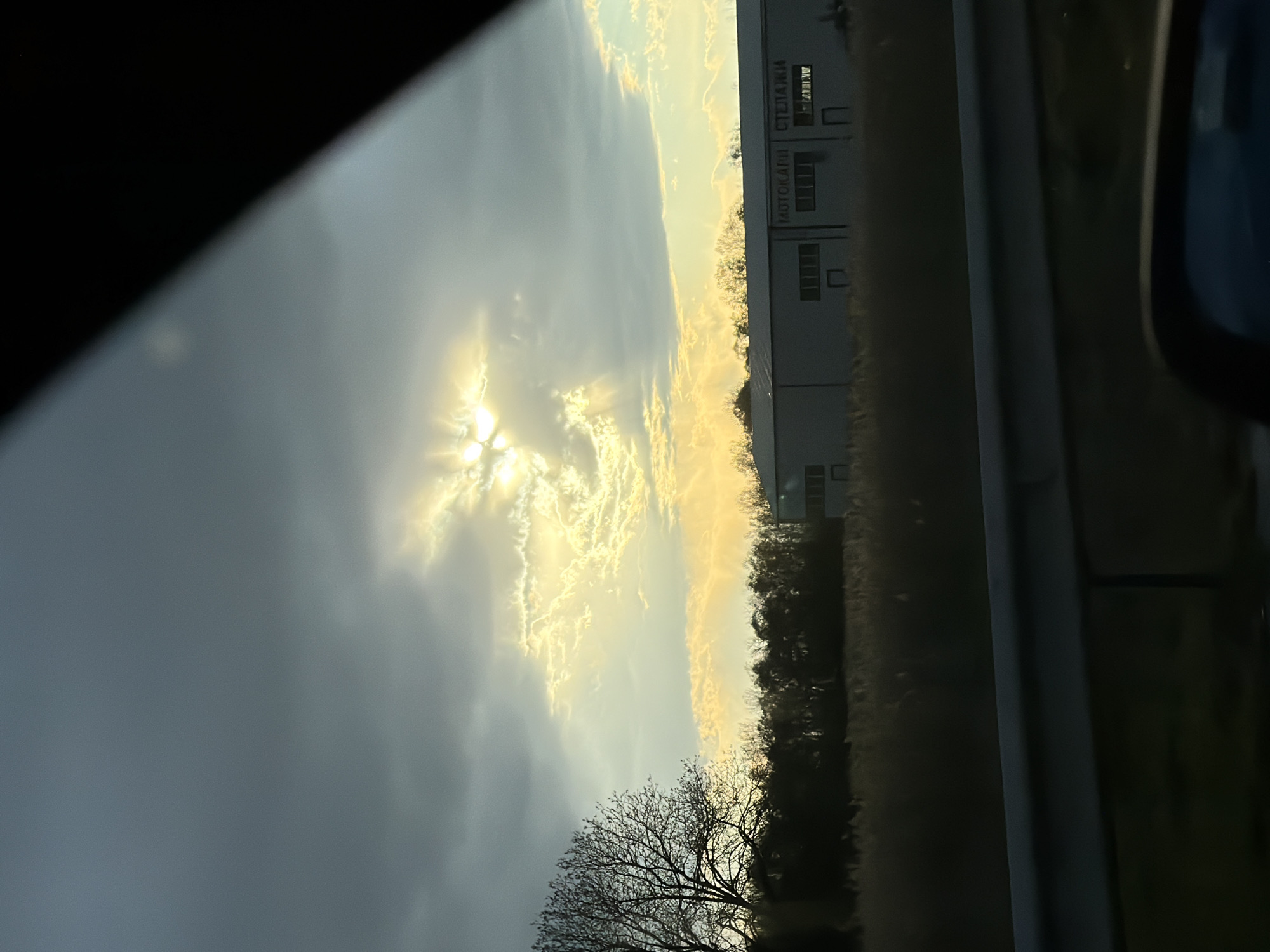 The image, taken from inside a vehicle, depicts a view towards the northeast, through the passenger window. The foreground shows the vehicle's window frame, partially obscuring the view.
The middle ground reveals a sky dominated by clouds, with the sun partially visible, creating a halo effect. Below the clouds, a line of trees separates the sky from a low-lying field. Adjacent to the field, a large industrial building, identifiable as "КОТОКАПИ СТЕЛАЖИ" ("KOTOKAPI Stelazhi") is visible. The text identifies the location as being in or near Plovdiv, Bulgaria, specifically the area where Kotokapi Stelazhi is located.
The background shows additional sky and cloud formations. There are no people visible in the image. The scene appears to be taken during the daytime, most likely in the early morning or late afternoon, given the angle and intensity of the sunlight. The overall environment is a mix of industrial and natural elements.