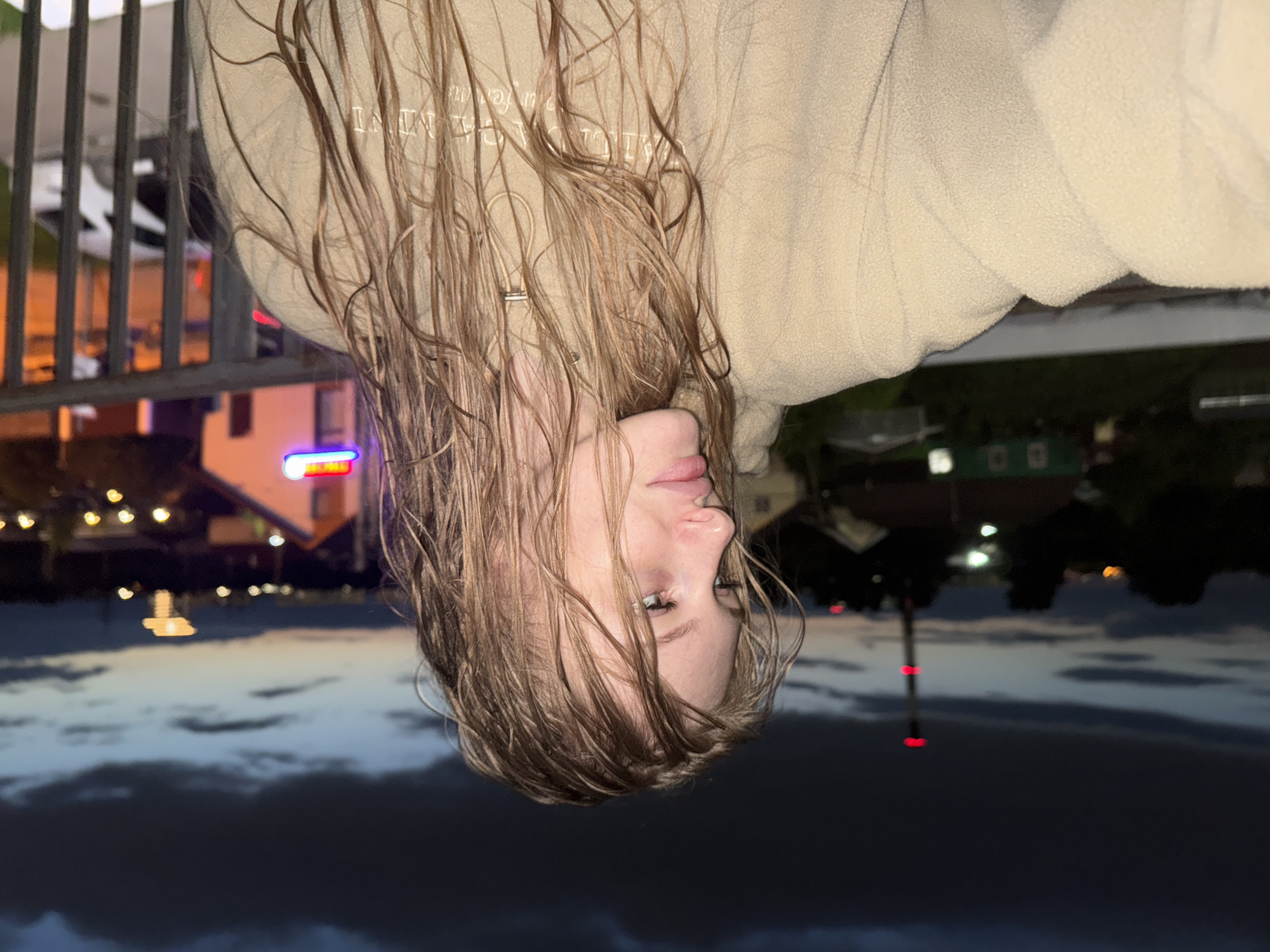 The image shows a young woman with wet blonde hair, wearing a light brown hoodie, standing on a balcony at dusk. She is looking to her left. In the background, a dimly lit cityscape is visible with houses, streetlights, and a tall industrial chimney with red lights. The location is identified as Kondratovo, Russia. A metal railing is in the foreground.