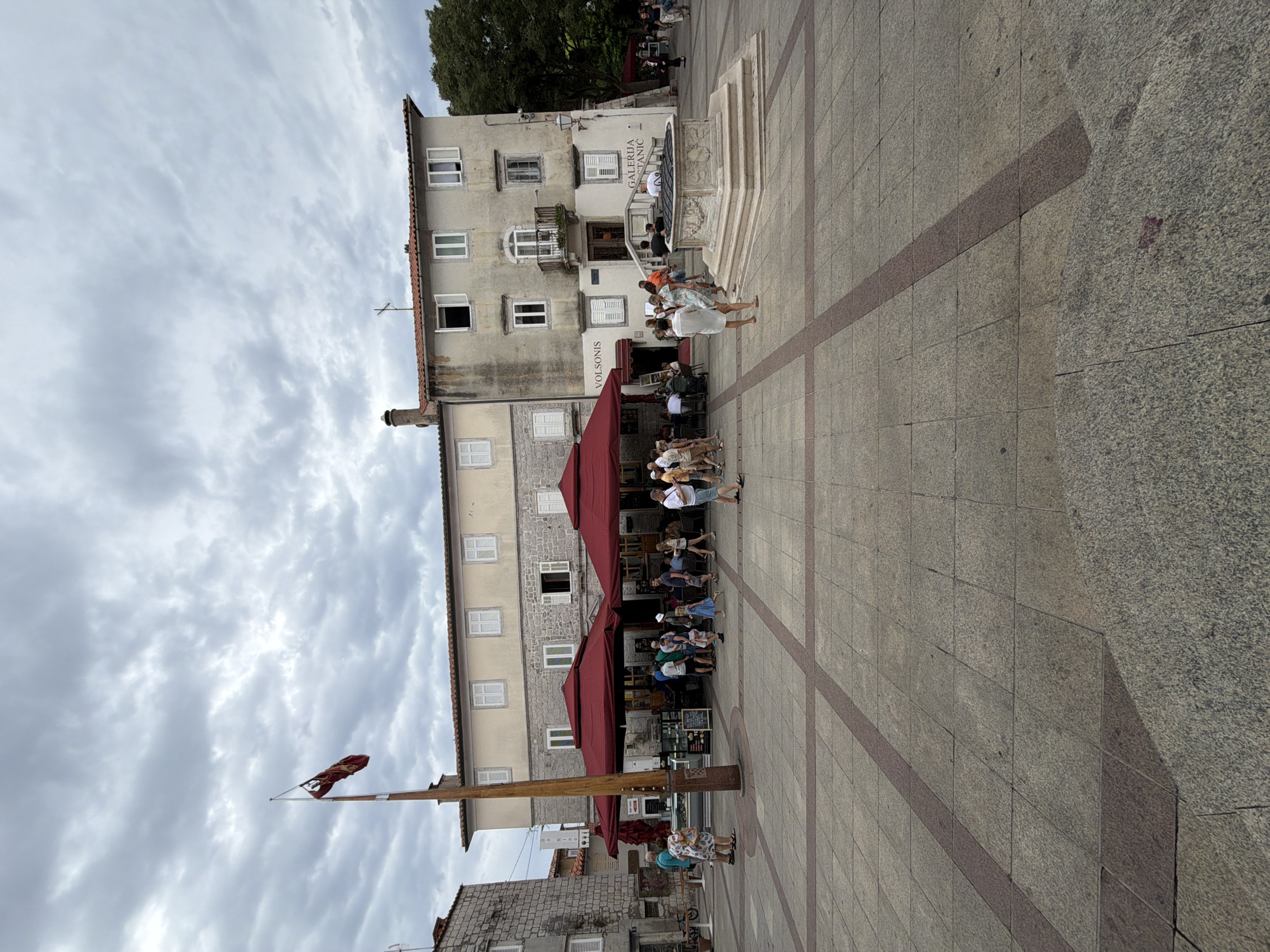 In the city of Krk, Croatia, an overcast sky looms over a paved town square. The square is bordered by a two-story building with a tan facade and white shutters, featuring multiple windows and a balcony on the second floor. To the left of this building, another stone building with similar windows stands. A tall wooden flagpole with a red and gold flag stands in the foreground. Underneath two large, red awnings, people are gathered at tables, likely at an outdoor cafe or restaurant. A stone well with intricate carvings is situated in the center of the square. Several people are walking around the square, some interacting with each other, while others are seated at the outdoor tables. The ground is paved with large square tiles, with red rectangular tiles forming a pattern. The scene depicts a typical day in a European town square, with people enjoying the outdoors amidst historical architecture.