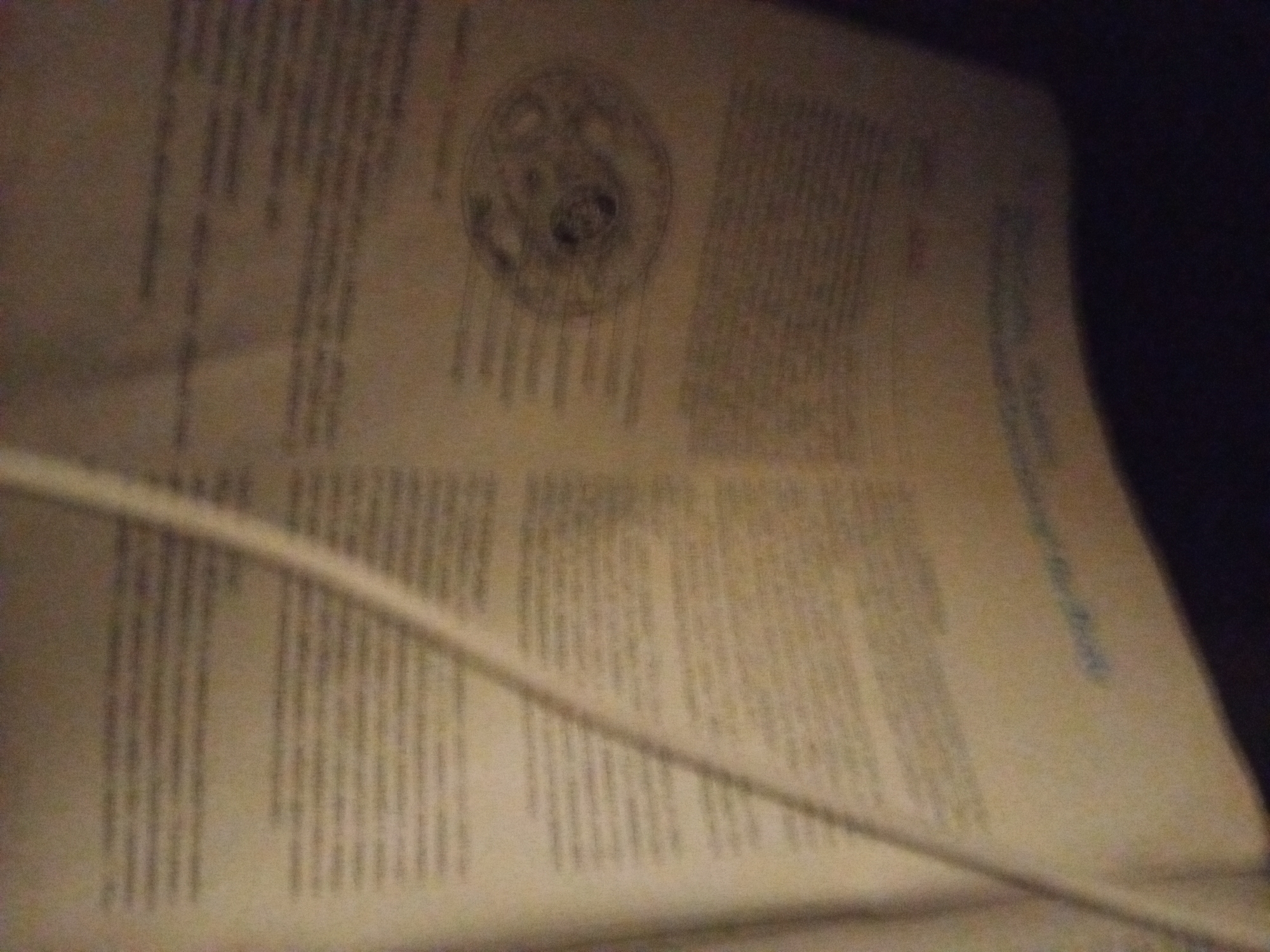 The image shows a close-up, angled view of what appears to be a page from a book or document, with text printed in columns. A thin, light-colored cable runs diagonally across the page, obscuring some of the text. In the upper left quadrant of the image, there is a circular diagram or illustration with several concentric rings and some darker interior details. The background is dark and out of focus, suggesting the photograph was taken in low light conditions. The paper appears aged and yellowed. There are no people or identifiable landmarks visible.
