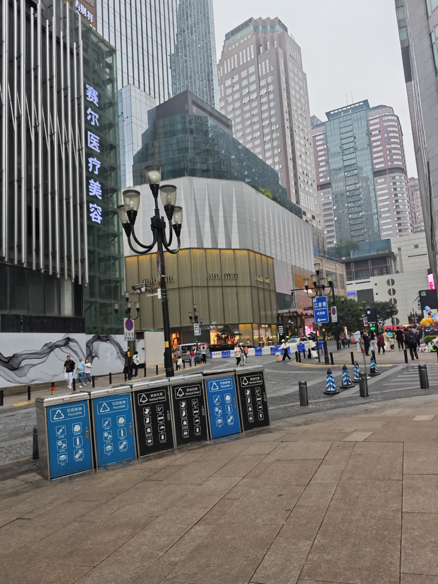 The photograph captures an urban street scene in Yuzhong District, Chongqing, China. In the foreground, a row of recycling and trash receptacles are prominently displayed, labeled in both Chinese and English as "Recyclable" and "Residual Waste," respectively.
In the background, people are walking on the sidewalks and crossing the street, suggesting a bustling urban environment. The architectural landscape is dominated by tall buildings, indicating a dense business district. One building features illuminated Chinese characters for “赛尔医疗美容”, denoting a medical aesthetic clinic. Streetlights with multiple lamp heads stand along the sidewalk.
Signage, including street names in Chinese such as 江家巷, adds to the authenticity of the location. Overall, the image depicts a typical street scene in a modern Chinese city, complete with waste management infrastructure and a busy urban atmosphere.
