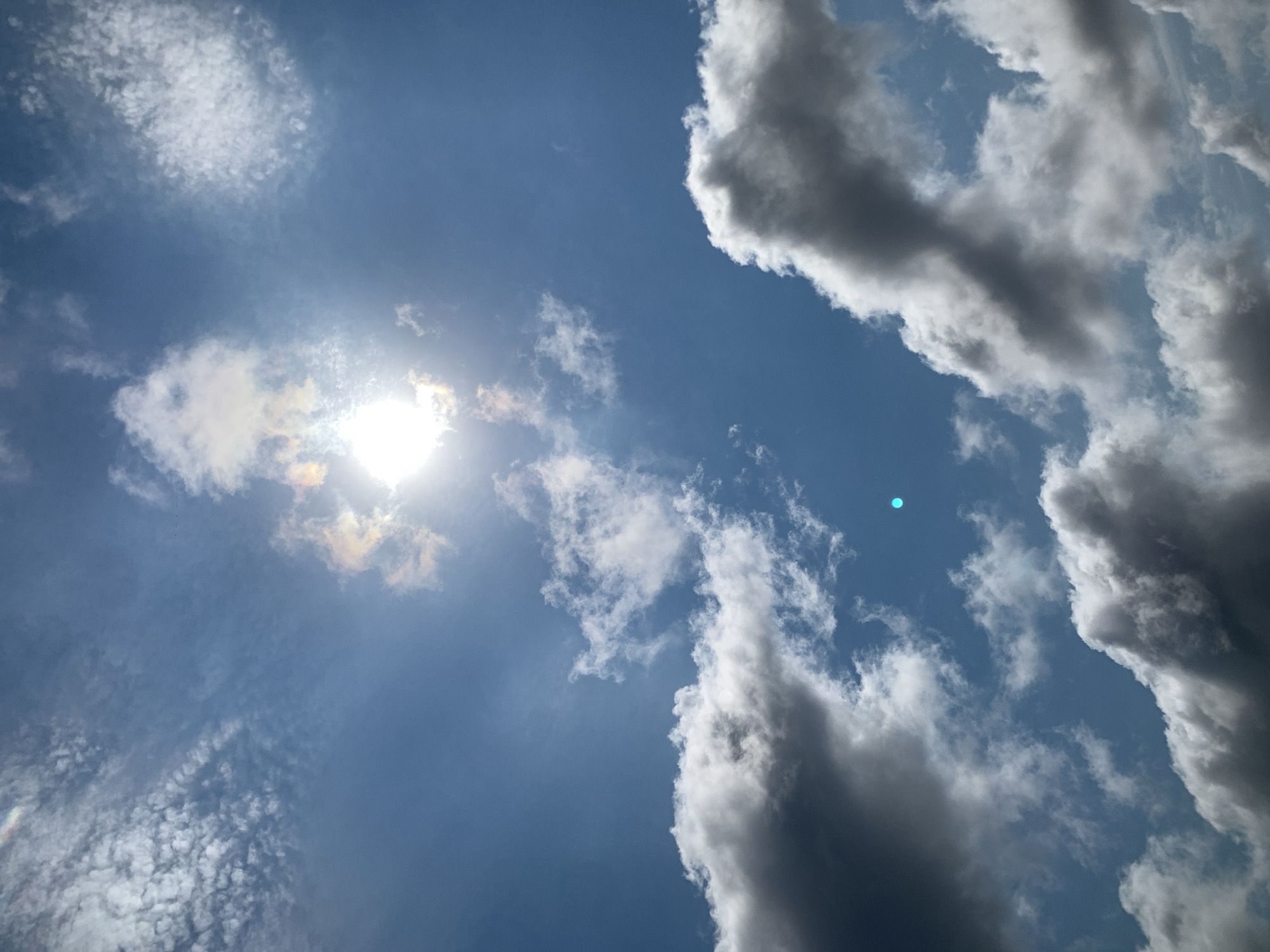 The image shows a bright sun with a halo effect, shining through a blue sky dotted with various clouds. The clouds range from wispy cirrus formations to larger, more dense cumulus clouds, some of which are shadowed. In the lower portion of the image, dark, dramatic clouds are visible, contrasting with the brighter sky above. A small, bright blue circular object is also present in the sky. The scene depicts a cloudy day with the sun breaking through, creating an atmospheric effect. The image's location is specified as Khazar Raion, Azerbaijan.