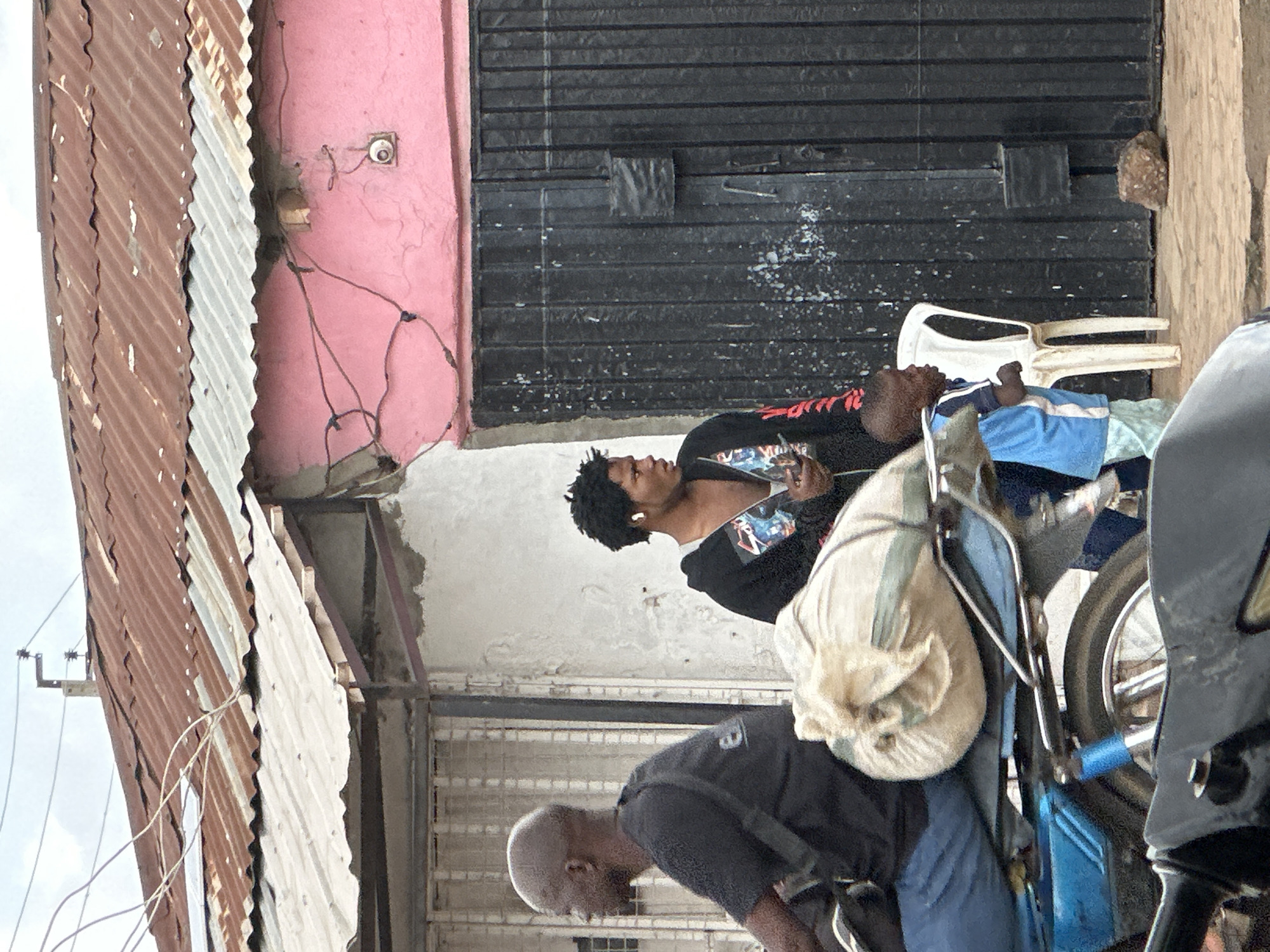 In Narayi, Nigeria, a motorcyclist wearing a black t-shirt and blue jeans is positioned on a blue motorcycle. A large, light-colored sack is secured on the back of the motorcycle behind him. To the right, a young man with dark curly hair wears earbuds and a black hoodie with a graphic design. He holds a phone in his right hand. A young child in a blue and white shirt stands near him. Behind them, a building features a pink-painted wall and a corrugated metal roof that appears weathered and rusted. A black metal gate is visible on the building's facade. The ground is paved with dirt and scattered with rocks. Electrical wires are strung overhead.