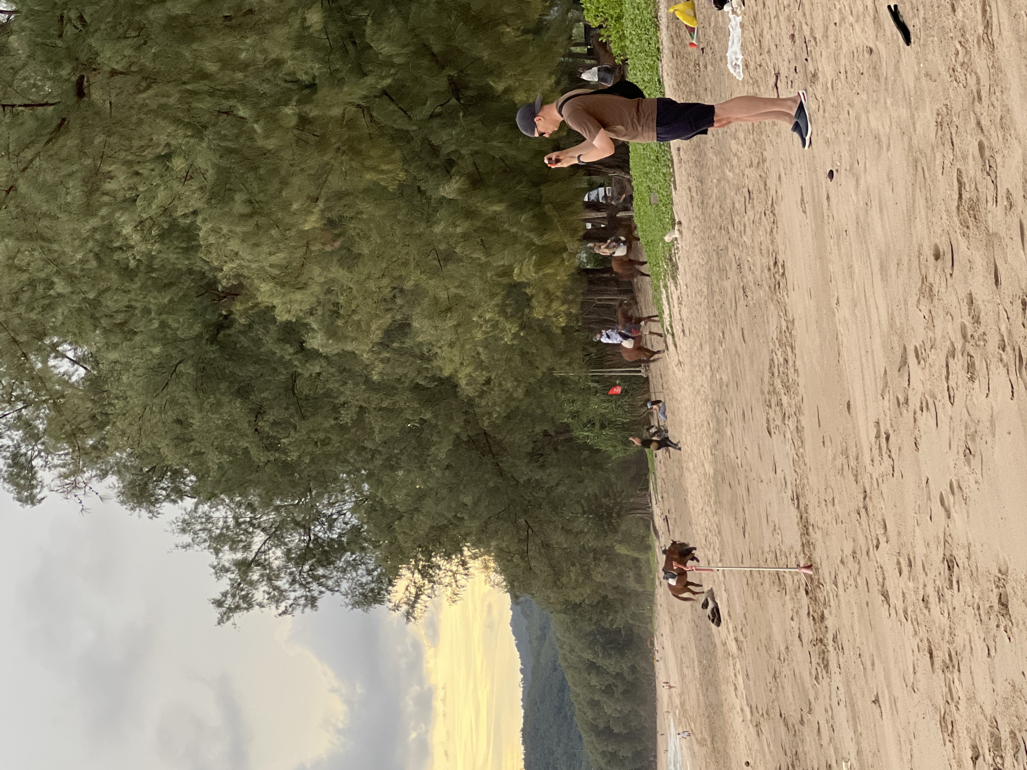 A man wearing a brown t-shirt, black shorts, a baseball cap, and slip-on shoes stands on a sandy beach, holding a phone to take a picture. He is facing towards the left of the frame, where several people are riding horses on the beach. A dense line of trees runs along the edge of the beach, and in the distance, a hazy line of hills is visible under a cloudy sky with a soft yellow glow. The sand is marked with footprints, and a few scattered objects, including a yellow bag and a white towel, are visible on the right side of the image.