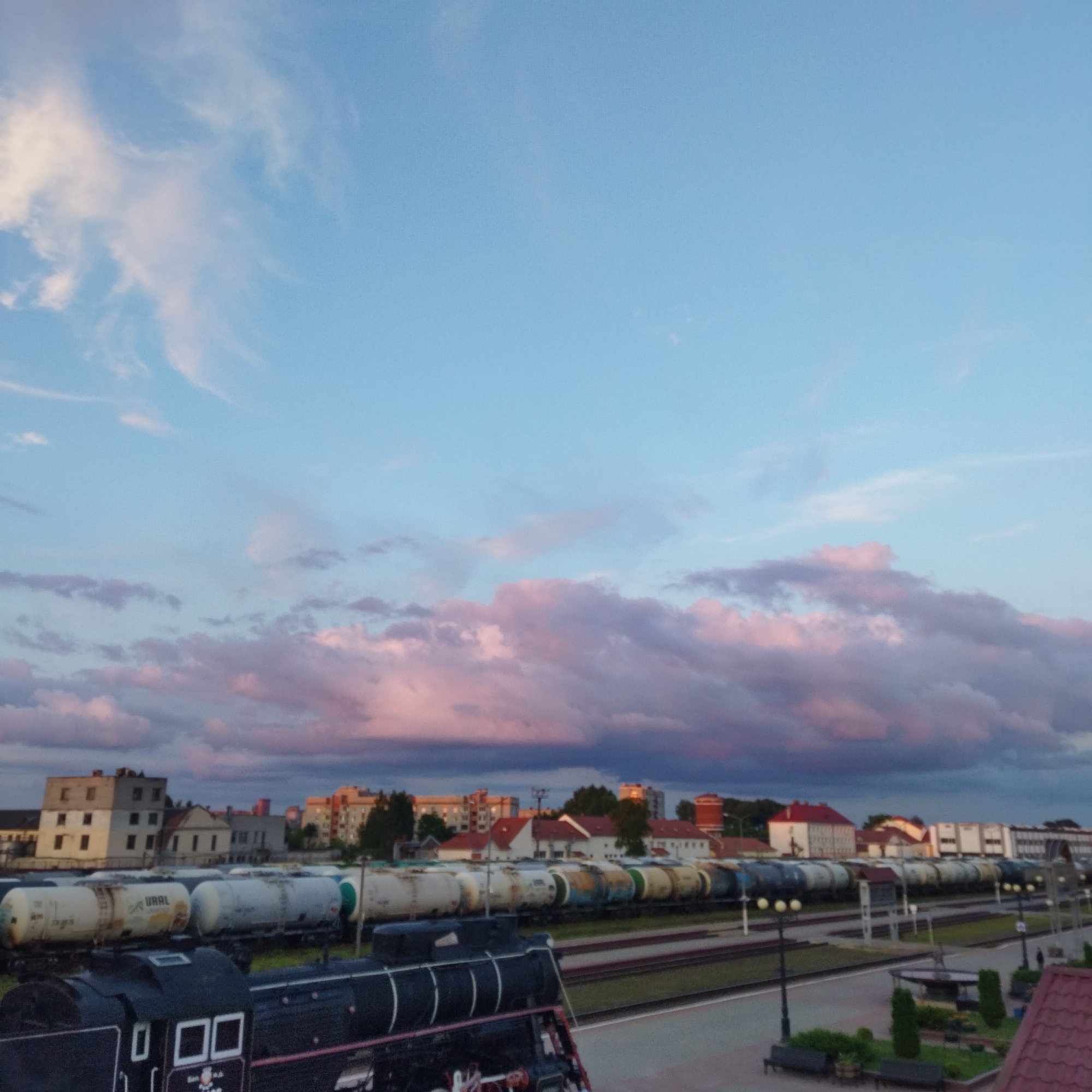The image shows a train yard in Polatsk, Belarus, with a vintage steam locomotive in the foreground and a line of freight cars behind it. In the background, buildings and trees are visible under a twilight sky with pink and blue clouds. The train cars appear to be oil or chemical tankers. A platform and streetlights suggest this is an active station.