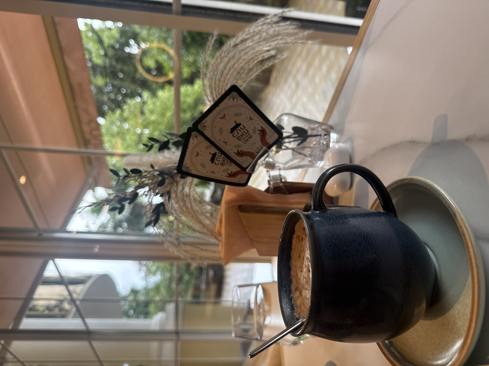 A dark blue mug filled with a frothy brown beverage, likely coffee or hot chocolate, rests on a light-colored saucer. A silver spoon is submerged in the drink. To the left of the mug is a glass of water with ice. Behind the mug and glass, a wooden napkin holder contains orange napkins. Two small, square cards with illustrations and text in an unfamiliar script are propped up in a clear glass vase filled with decorative clear beads and dried foliage. The scene is set on a white marble-patterned table. In the background, large windows offer a view of a building with white columns and balconies, as well as lush green trees and foliage outside. The lighting suggests an indoor setting, possibly a cafe or restaurant. The overall impression is one of a tranquil moment, perhaps a break at a cafe in Sochi, Russia.