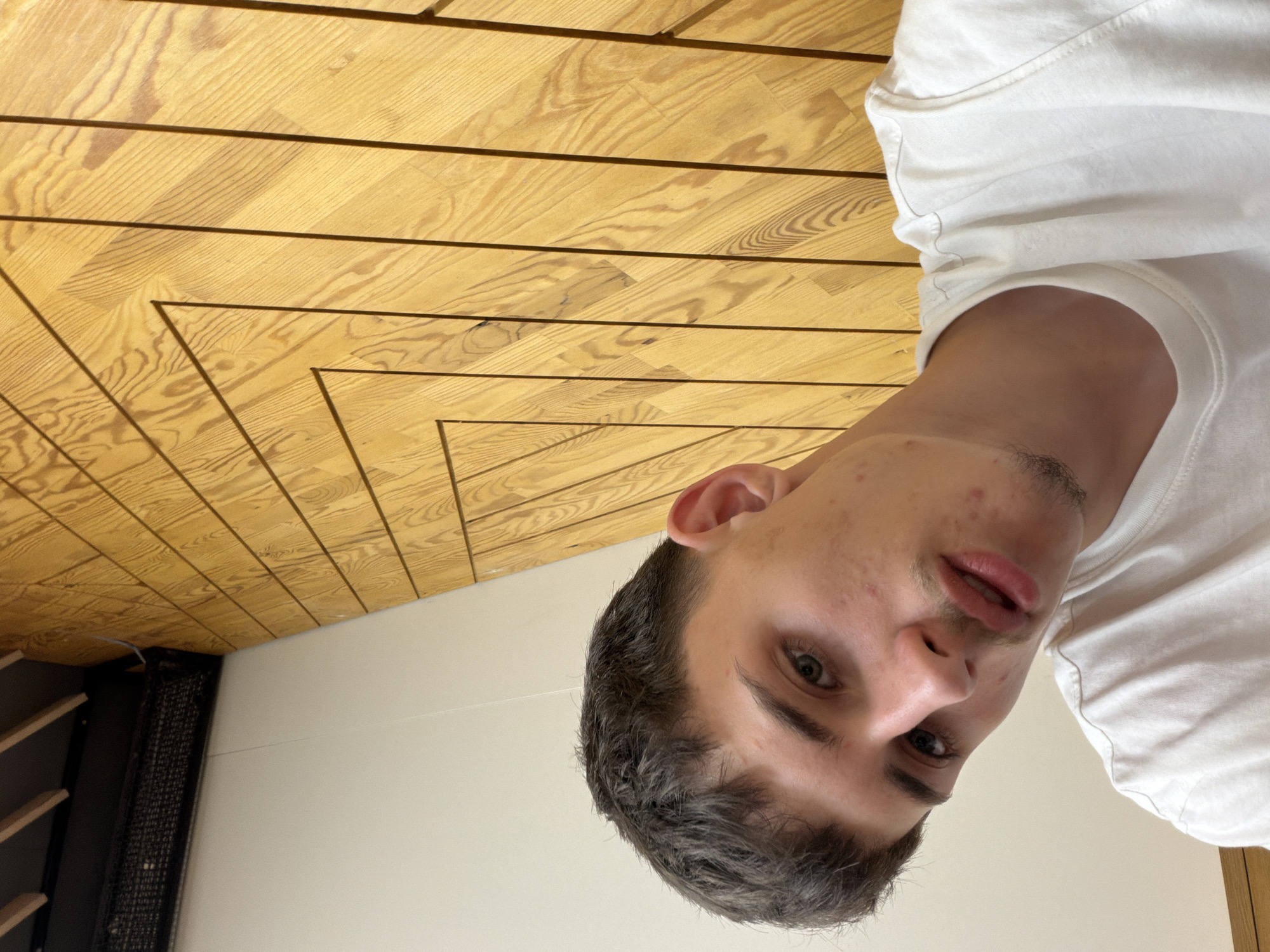 The image shows a young man taking a selfie in front of a wooden wall with a geometric pattern. The man is wearing a white t-shirt and has short, dark hair. His face is turned towards the camera, and he appears to be looking directly at it. He has light-colored eyes and some acne on his face. The wooden wall is made of light-colored wood and is arranged in a series of concentric squares. The lighting in the image is bright, suggesting it was taken during the day. The overall impression is a casual, personal snapshot. The location is not specified in the image, and therefore it cannot be determined if it is in Plovdiv, Bulgaria.
