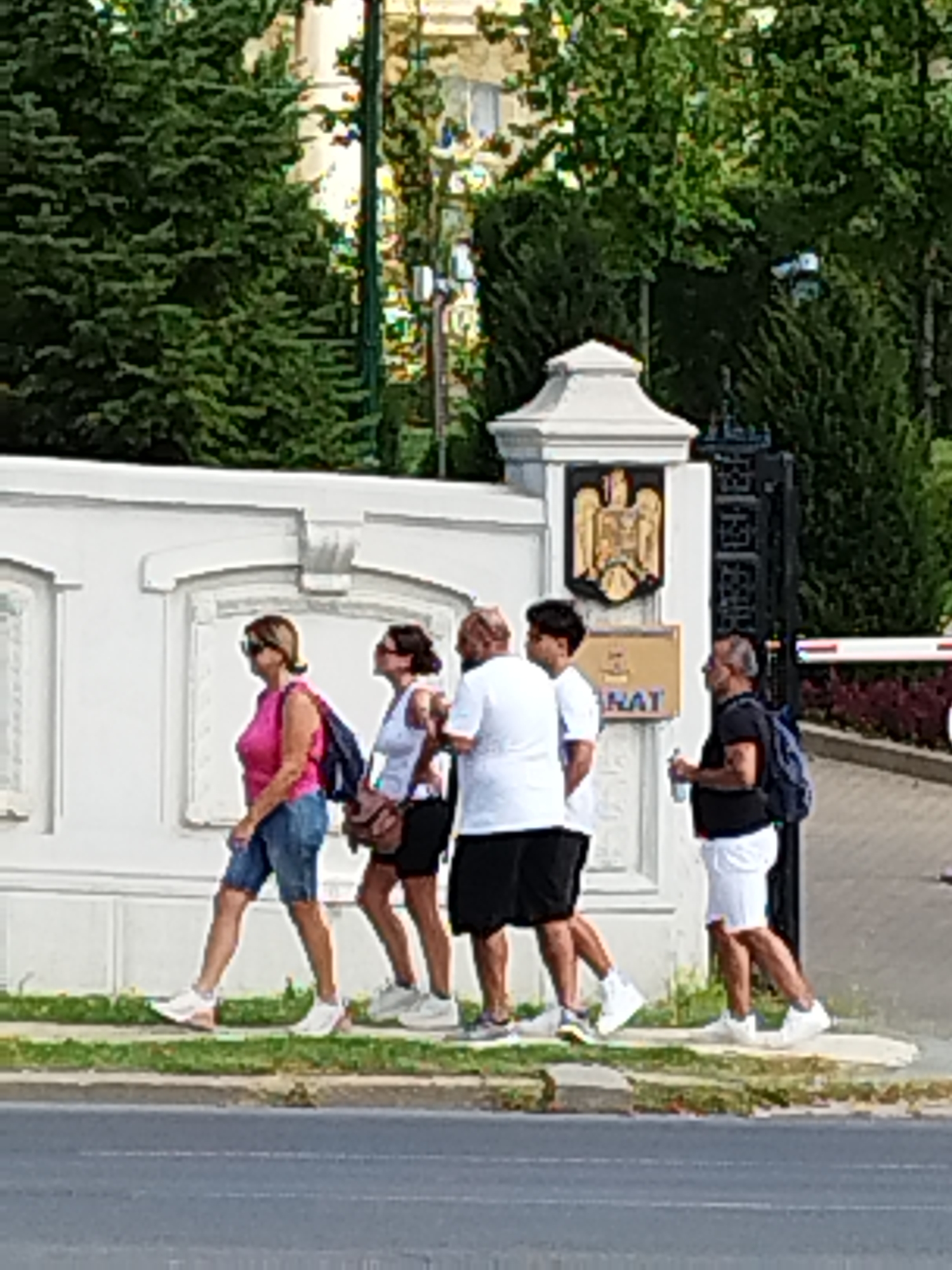 The image depicts a group of five individuals walking on a sidewalk in front of a white wall with arched insets. A dark metal gate is visible to the right, with a red and white barrier gate beyond it. A coat of arms is displayed on a white pillar to the left of the gate. The individuals include three men and two women, all dressed in casual attire. They are walking from left to right, with the woman in front wearing a pink shirt and denim shorts. The scene appears to be located in Bucharest, Romania, likely near a government building, given the coat of arms. The environment suggests a daytime setting with lush green trees in the background.