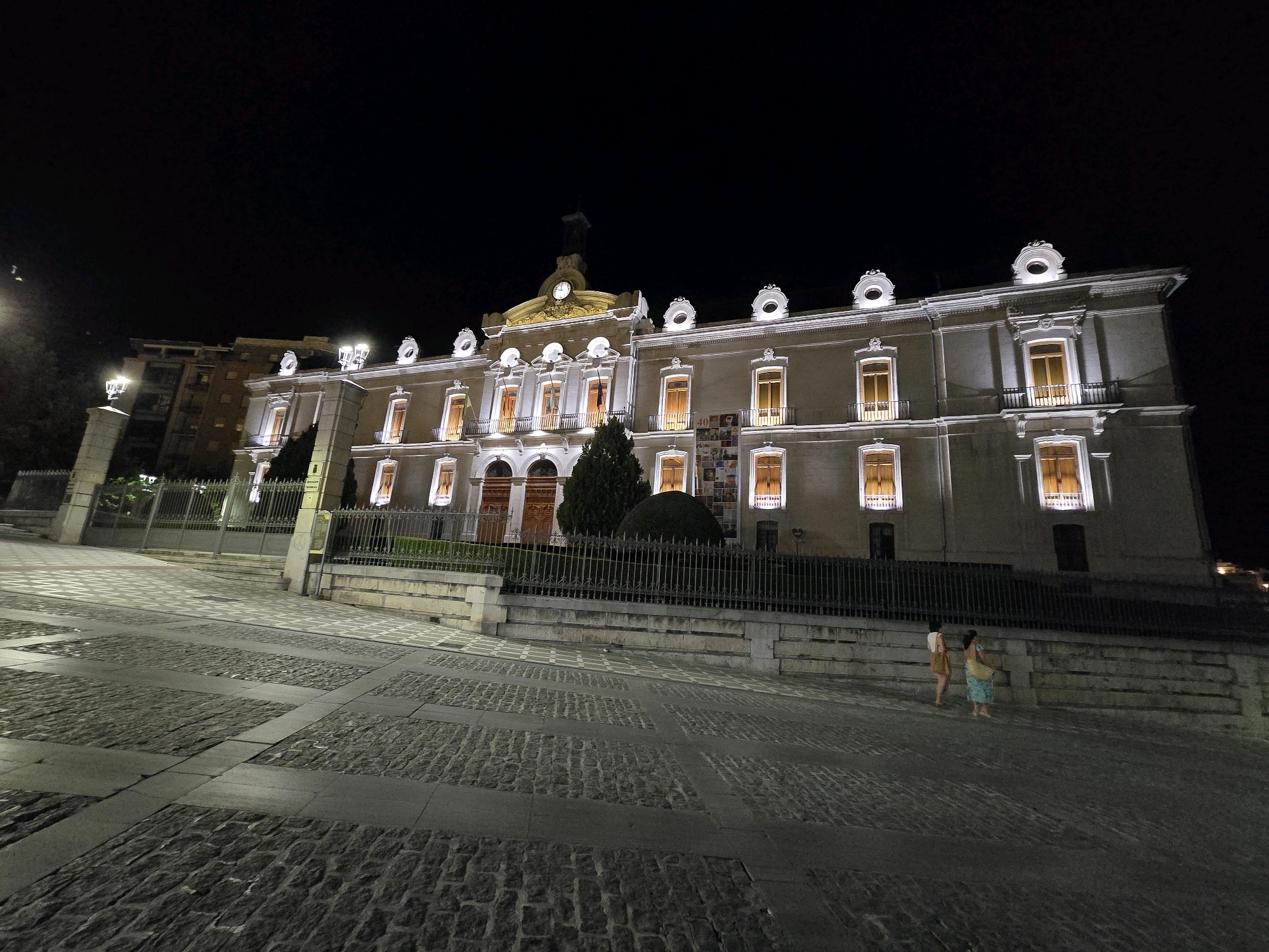 The image captures the Palacio de Congresos y Exposiciones in Jaén, Spain, at night. The imposing building, illuminated by interior lights emanating from its many windows, is the central subject. The facade features a classical architectural style with ornate details around the windows and along the roofline. A clock is visible on a pediment above the central entrance.
In the foreground, a cobblestone plaza stretches out, with a distinct pattern of lighter and darker stones forming geometric designs. A low, ornamental metal fence separates the plaza from the building's grounds, which are lined with manicured hedges and trees.
Two individuals are present on the plaza, walking away from the camera. The person on the left appears to be wearing a light-colored top and shorts. The person on the right is wearing a patterned dress and carrying a bag over their shoulder. They are walking on the cobblestone surface. The overall scene is dimly lit by artificial lights, creating a dramatic ambiance typical of a nighttime urban environment. The sky is dark and featureless, indicating the absence of moonlight or visible stars.