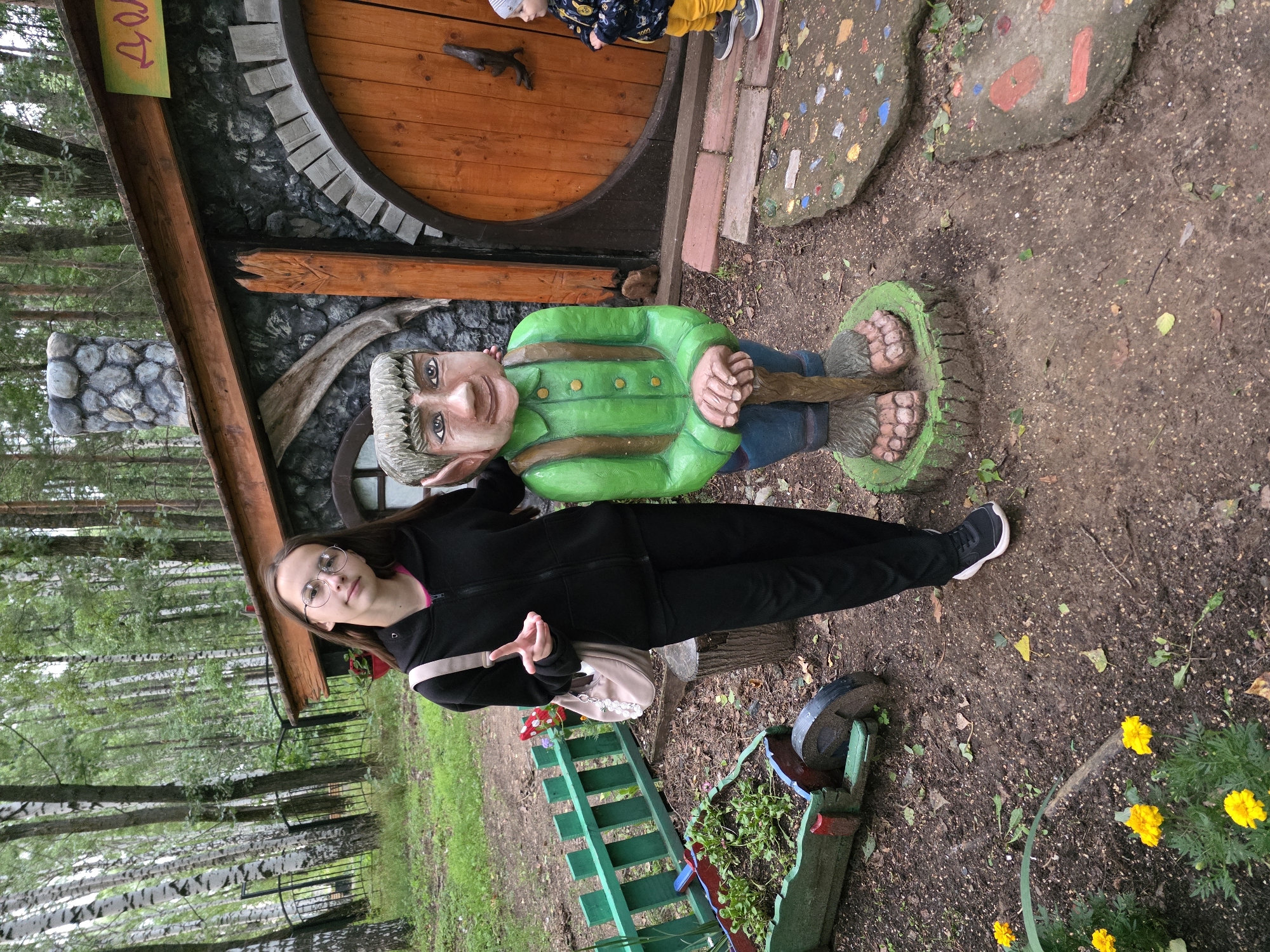 The image shows a young woman with long brown hair, wearing glasses, a black zip-up hoodie, and black pants, standing next to a wooden statue of a gnome. The gnome statue is painted green and blue, with brown suspenders and a brown staff. Behind them is a rustic-looking house with a round wooden door and a stone chimney. The setting appears to be an outdoor park or garden area, with trees and greenery in the background, and a small green fence and a wooden cart with plants in the foreground. A young child in a blue jacket and yellow pants is visible to the right of the house. The scene is likely in the city of Chaikovsky, Russia, as indicated by the context.