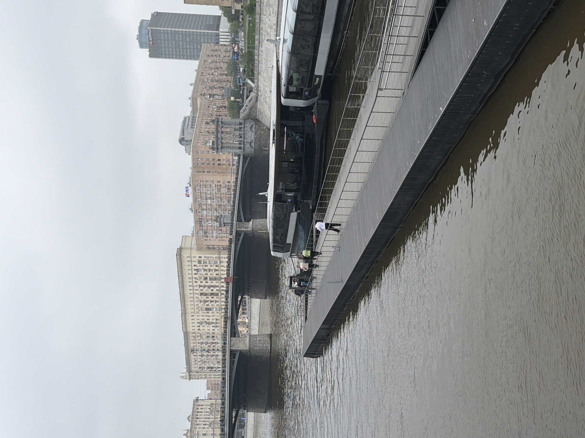The image shows a watercraft docked on the Moscow River in Moscow, Russia. A bridge spans the river in the background, flanked by numerous high-rise buildings. Several individuals stand on the dock or are boarding the watercraft. The weather appears overcast, with a gray sky. The river water has a muddy appearance.