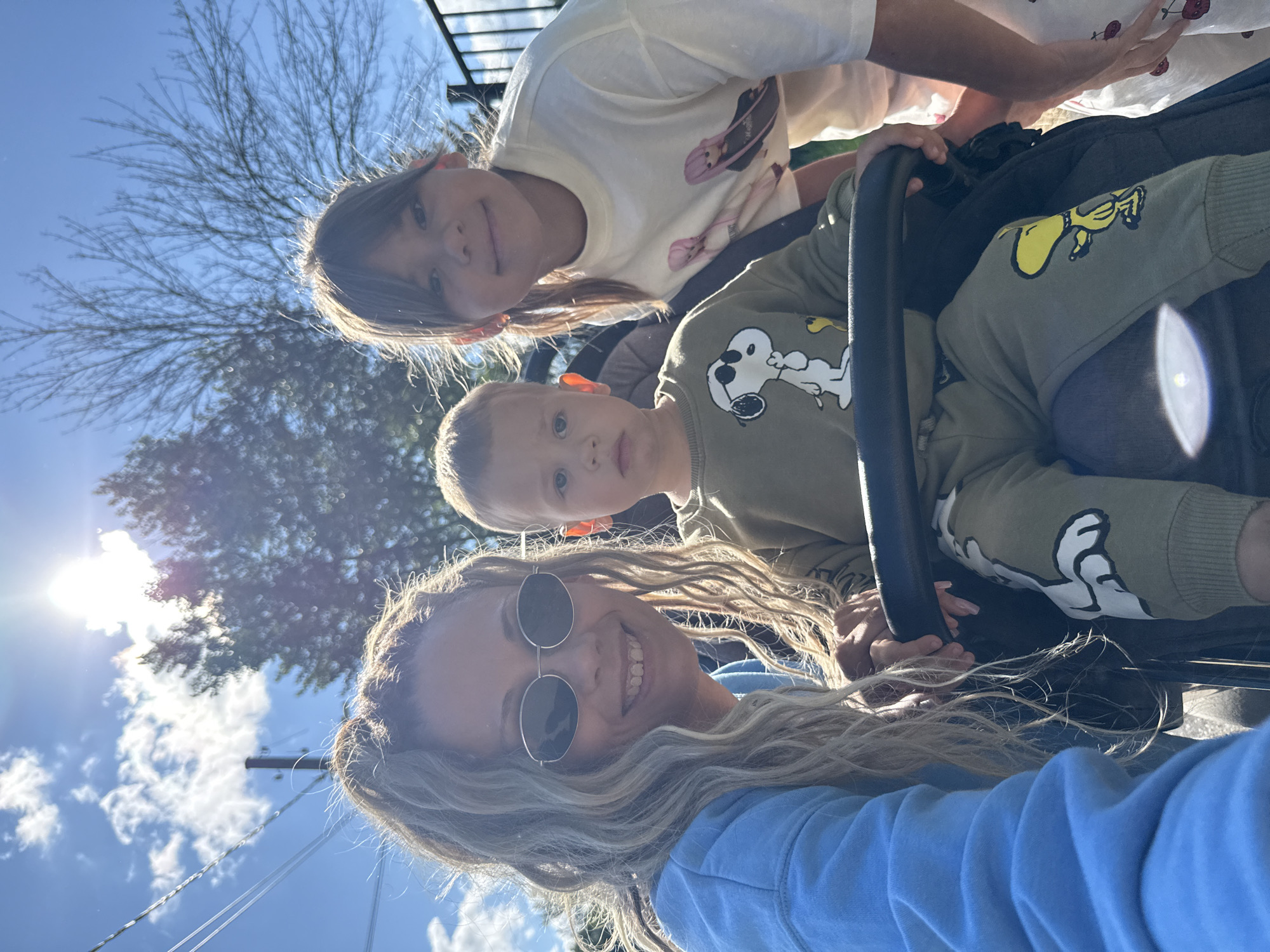 The image captures a selfie taken outdoors on a sunny day, with a bright sky and scattered clouds. The foreground features a woman wearing sunglasses and a blue hoodie, smiling at the camera. To her right, a young boy in a stroller is seated, wearing a green sweatshirt with Snoopy on it and matching pants. Behind the boy, a girl with her hair in a ponytail, wearing a cream-colored t-shirt with cartoon characters, is leaning forward and smiling. The background shows trees, a utility pole with wires, and a dark fence. The scene appears to be in a residential or park area. There is no visual information that suggests the location is in Rusanovo, Russia.