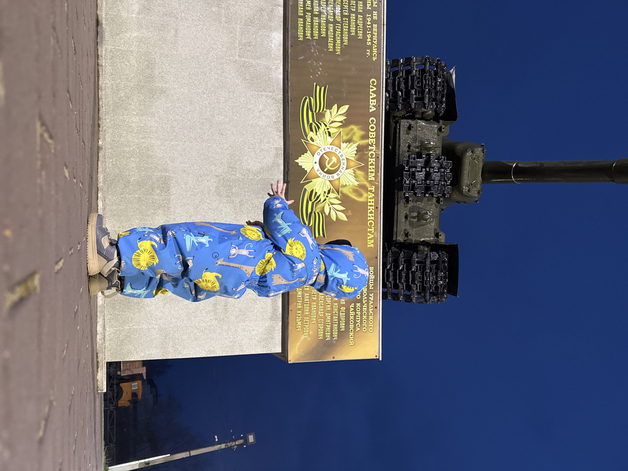 A young child, dressed in a blue full-body suit patterned with yellow lions and other animals, stands on a light-colored concrete path. The child, wearing grey and beige shoes, is extending their right arm and hand towards a monument. The monument consists of a light-colored concrete base, upon which a dark bronze-colored plaque is affixed.
The plaque features Russian text, a golden star emblem (likely the Soviet Order of the Patriotic War), and several yellow and black striped ribbons. Above the emblem, the text reads "СЛАВА СОВЕТСКИМ ТАНКИСТАМ" (Glory to Soviet Tankers). The emblem itself includes the words "ОТЕЧЕСТВЕННАЯ ВОЙНА" (Patriotic War). Surrounding text lists names of individuals who "НЕ ВЕРНУЛИСЬ" (did not return) from "1941-1945 гг." (1941-1945 years), identifying them as "БОЙЦЫ УРАЛЬСКОГО ДОБРОВОЛЬЧЕСКОГО КОРПУСА" (Fighters of the Ural Volunteer Corps), with the name "ЧАЙКОВСКИЙ" also visible.
Behind the plaque and integrated into the monument is a dark green tank, with its gun barrel pointing upwards and to the right. Sections of its tracks are visible, mounted as part of the display. The scene is set outdoors under a deep, clear blue sky, suggesting either dusk or night, illuminated by artificial light sources not visible in the frame. The child appears curious or playful, reaching out towards the memorial dedicated to Soviet tank crews from World War II, specifically mentioning the Ural Volunteer Corps, aligning with the given location context of Chaikovsky, Russia.