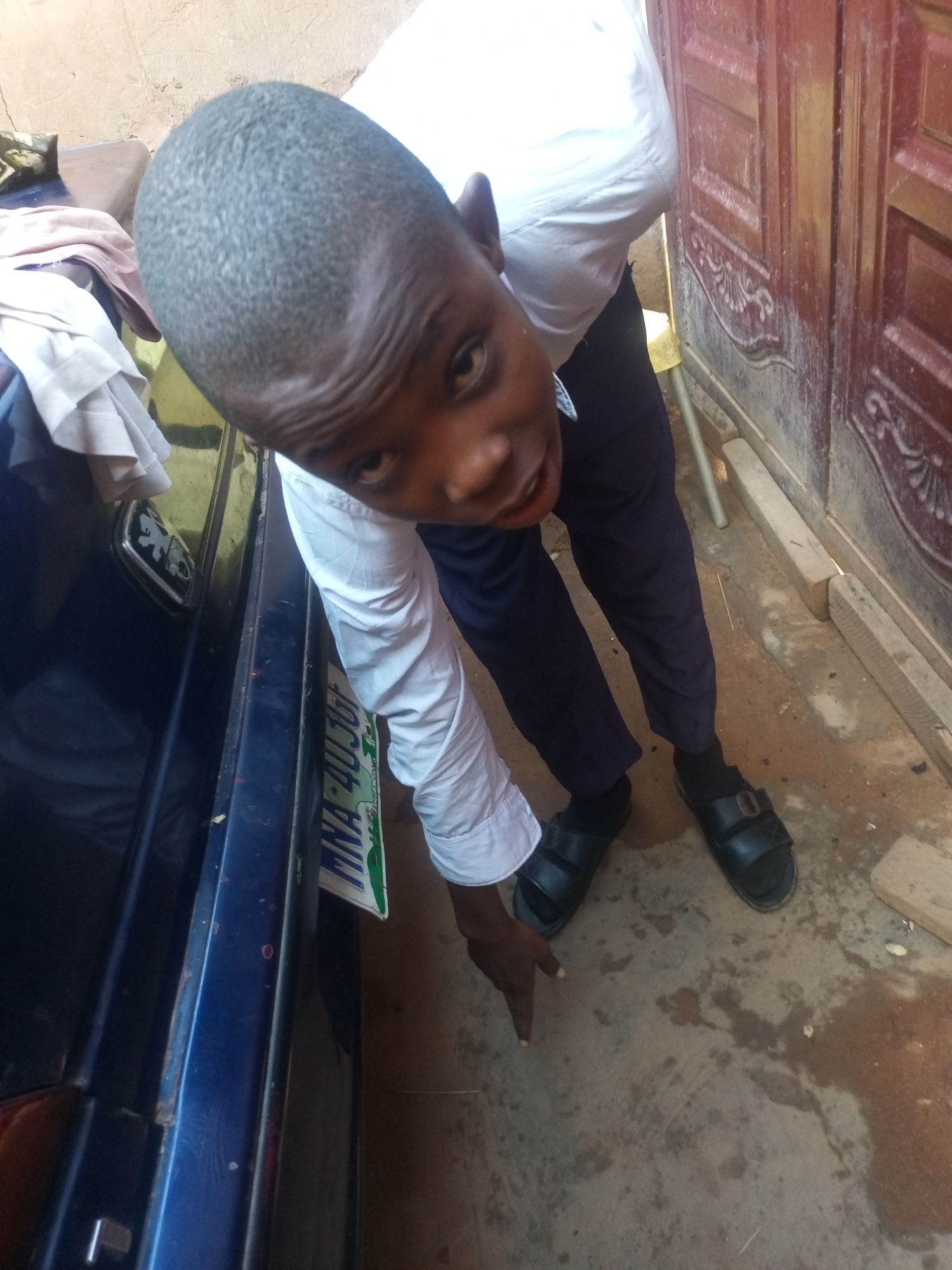 A boy, estimated to be between 8-10 years old, with a buzz cut and dark skin, wearing a white collared shirt and dark blue trousers, is leaning forward towards the viewer. He is wearing black sandals. His right hand is extended downwards, pointing towards the ground. He is standing next to a dark blue vehicle. A license plate is visible on the vehicle, with the text "MNA-40JU9" and green lettering. The ground is a mixture of concrete and dirt, with some wet patches. In the background, a wooden door with red paint and decorative carvings is visible. The scene appears to be an outdoor environment, possibly in a residential area of Tempure, Nigeria, given the visible architecture and the presence of the vehicle.