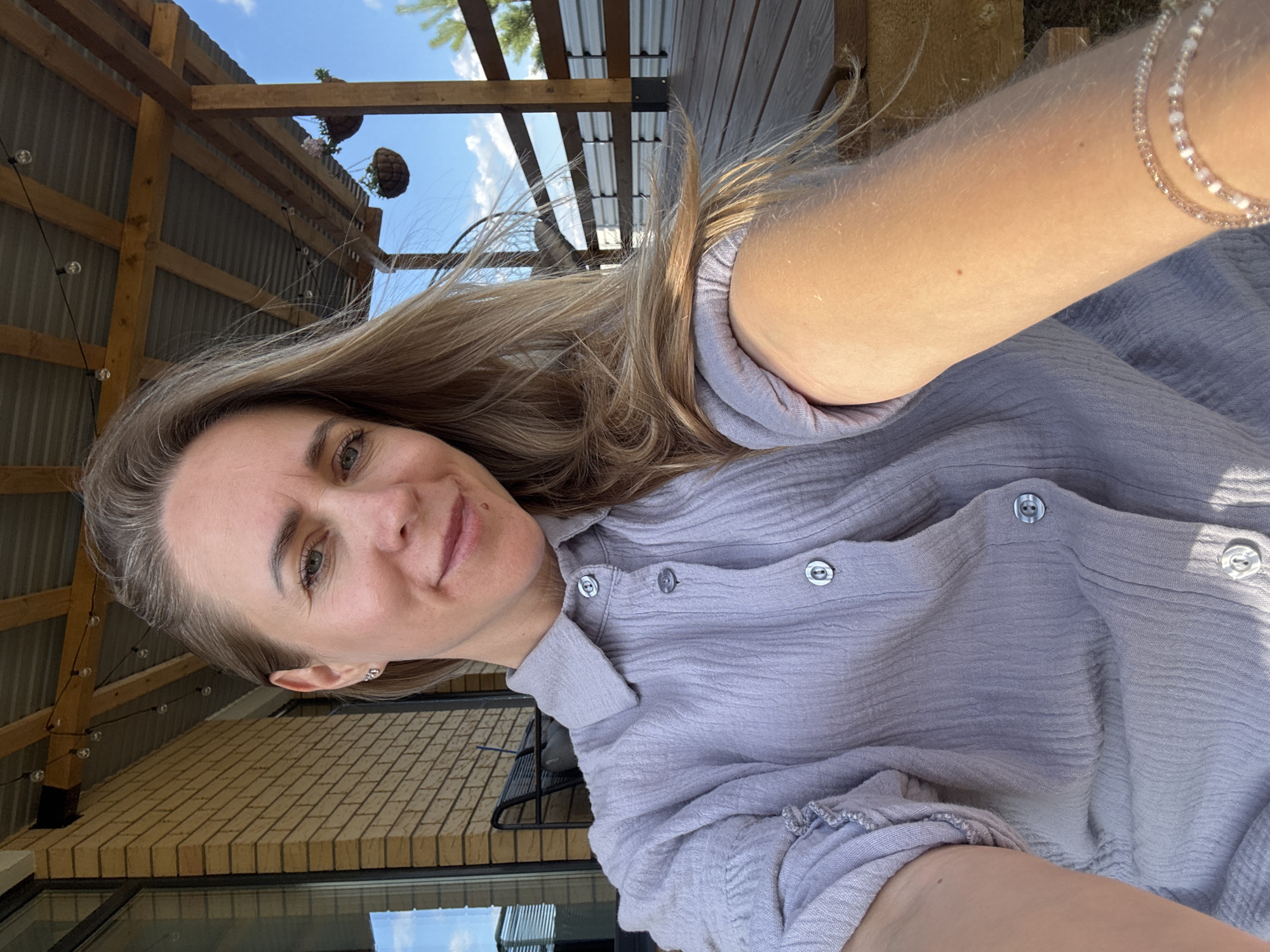 This is a vertical, selfie-style photograph of a woman with long blonde hair, smiling at the camera. She is wearing a light purple, textured button-up shirt. The setting appears to be outdoors, under a wooden structure with a corrugated metal roof. There are hanging plants and string lights visible above and behind her. Sunlight is casting shadows, suggesting it is daytime. The background also shows a brick building. The woman is wearing a beaded bracelet on her right arm. Her expression conveys happiness and relaxation. There is no visible text in the image.