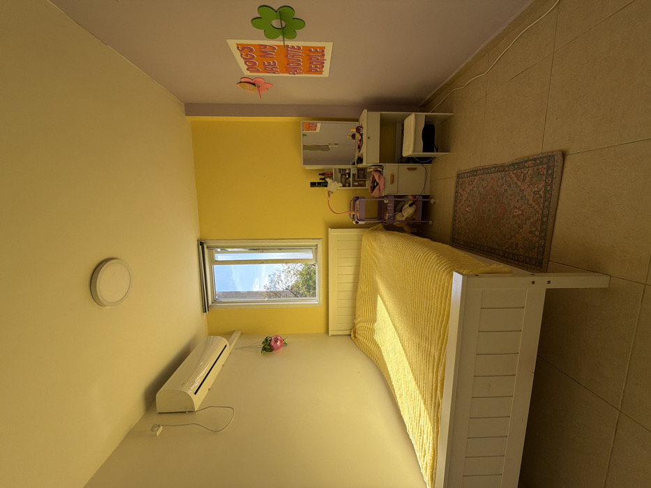 This image displays an interior view of a residential bedroom, specified to be located in the city of Tel-Aviv, Israel. The room features a multi-colored wall scheme: the left wall is light cream, the back wall with the window is bright yellow, and the right wall is light purple.
A white wooden single bed with a slatted headboard and footboard occupies the left side of the room. It is covered with a bright yellow, textured bedspread. Natural sunlight enters through a rectangular white-framed window on the yellow wall, casting a bright rectangular illumination pattern across the bedspread. Through the partially open window, blue sky and green tree foliage are visible.
Mounted high on the light cream wall, above the bed's head area, is a white split-system air conditioning unit with a visible power cord. Below it, a small pink heart-shaped hanging planter holds a green plant.
In the corner formed by the yellow and purple walls, a white wooden dressing table with an attached rectangular mirror is positioned. Various cosmetics, bottles, and a pink flexible-arm desk lamp are arranged on its surface. A small white, backless stool is tucked underneath the dressing table. Adjacent to the dressing table, against the yellow wall, is a light purple three-tier rolling cart containing miscellaneous items, including what appears to be a hair dryer.
The light purple wall features a white poster with orange text that reads "DOGS ARE MY FAVOURITE PEOPLE," flanked by a pink butterfly-shaped cutout to its left and a green flower-shaped cutout to its right.
The floor is covered with large, light beige/tan ceramic tiles. A small, rectangular, patterned rug with a traditional design (featuring reds, blues, and greens) lies on the floor between the bed and the dressing table. A circular white flush-mount ceiling light fixture is centered on the light cream-colored ceiling. The room is devoid of any individuals or active subjects.