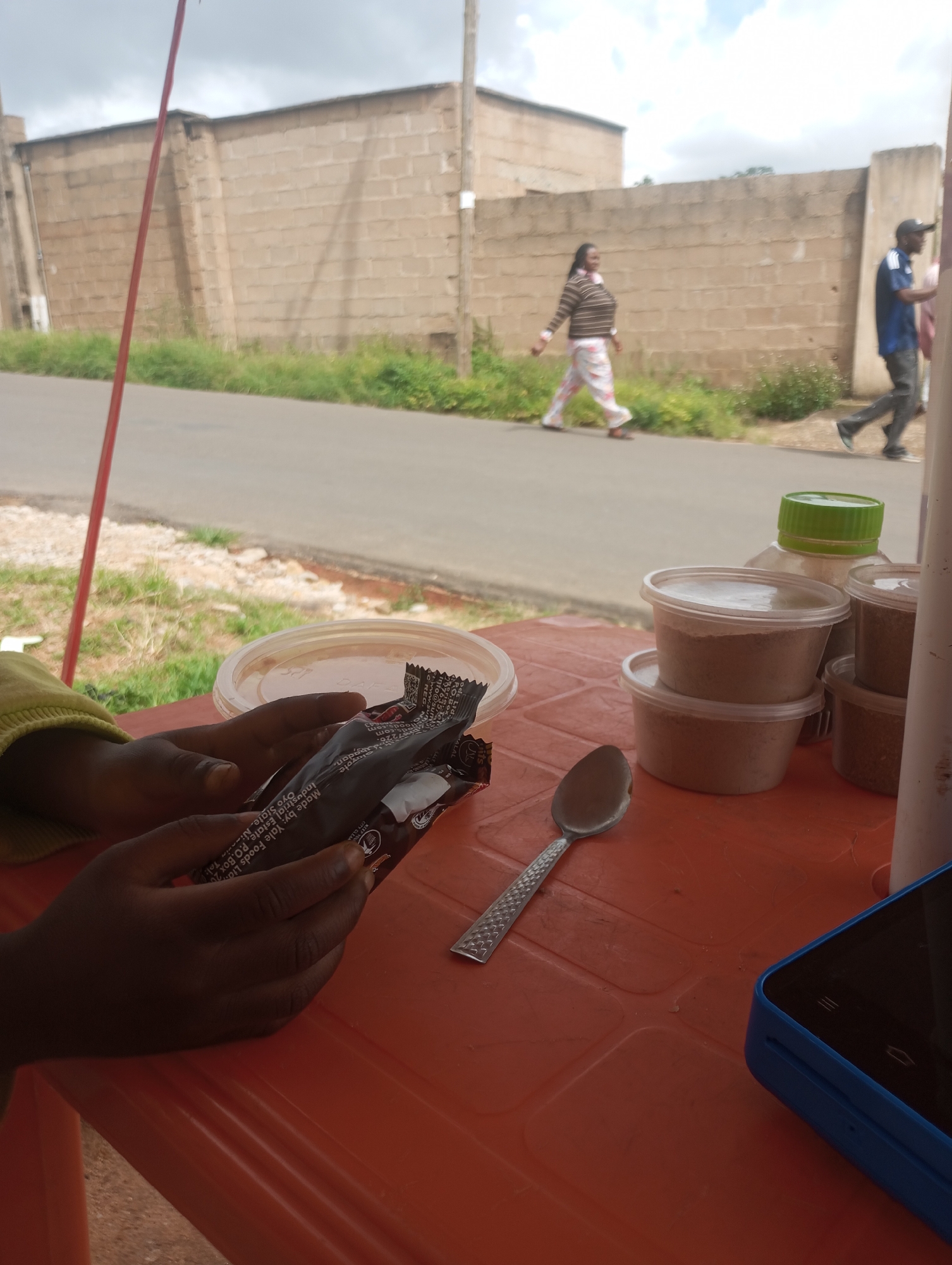 A medium shot captures a person's hands holding a dark-colored snack bar with text in English and what appears to be Arabic script on an orange plastic table. In the foreground, a silver spoon rests on the table, and a blue-cased smartphone is partially visible to the right. In the background, several plastic containers filled with a light brown substance are stacked, and a large green-capped bottle stands behind them. Beyond the table, a paved road runs parallel to a low brick wall, with green vegetation on the other side. Two individuals are walking on the road, one a woman in a striped sweater and patterned pants, and the other a man wearing a blue shirt and a baseball cap. The sky is overcast with scattered clouds. The scene is set in the city of Jos, Nigeria, indicated by the product labeling on the snack bar, which reads "Made by: Yale Foods Ltd, Industrial Estate, P.O. Box 2029, Gwagwalada, Oyo State, Nigeria." This implies the snack bar was manufactured in Nigeria, and the surrounding environment, including the brick wall and the presence of individuals of Nigerian ethnicity, is consistent with a location in Jos.