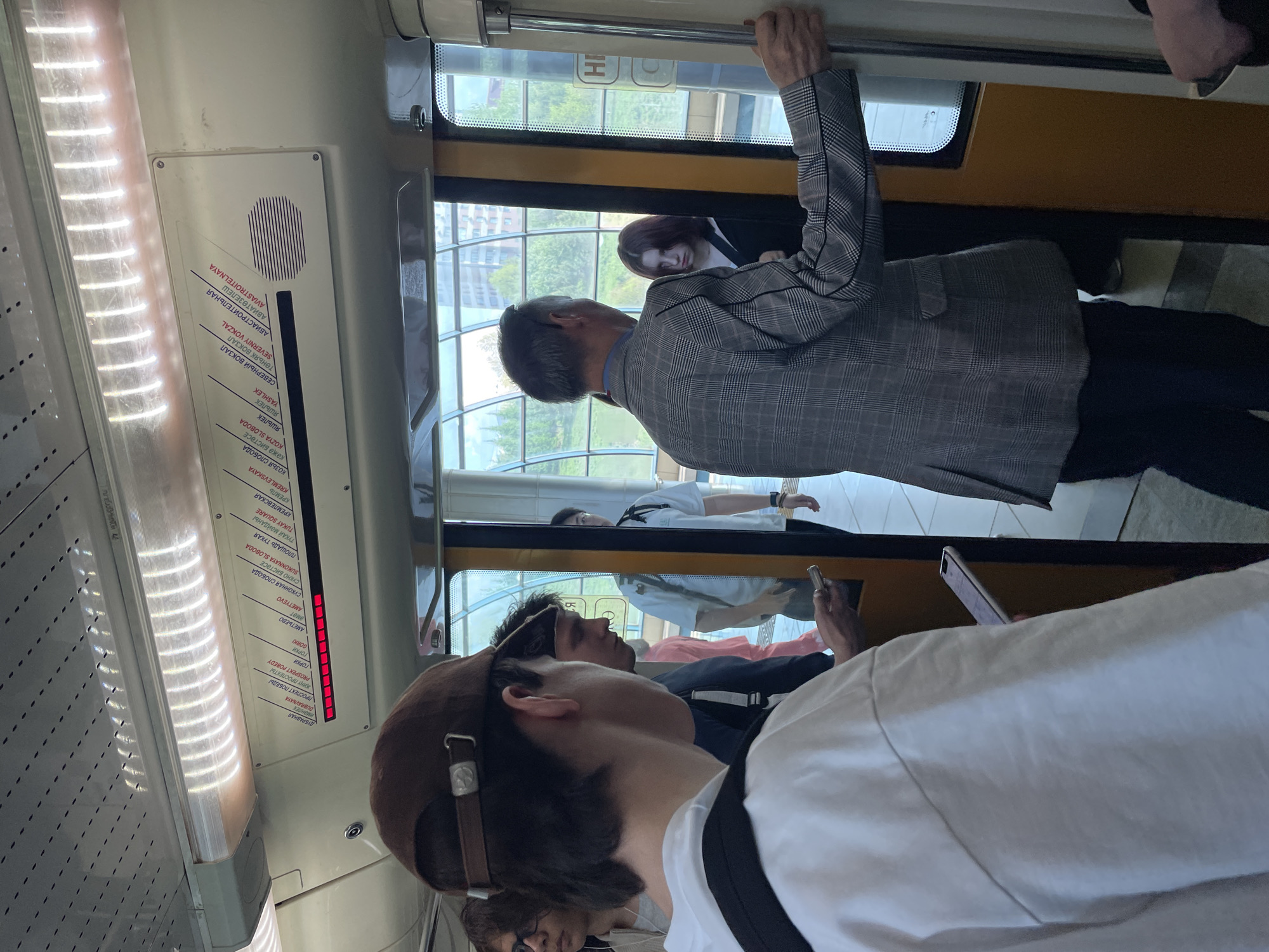 Here is a detailed description of the image:
**Scene:**
The image appears to be taken inside a monorail or elevated train in Kazan, Russia. The angle is slightly unusual, with the camera tilted, giving a dynamic perspective.
**People:**
Several individuals are visible in the train car:
* **Person in a Plaid Blazer:** A man is standing, holding onto a handrail. He is wearing a grey plaid blazer and dark trousers. He appears to be middle-aged or older and is looking out the window.
* **Person in the Foreground:** The closest person to the camera is wearing a brown cap and a white shirt with a black strap.
* **Other Passengers:** Other passengers are visible in the background, some looking at their phones.
**Objects:**
* **Train Interior:** The interior of the train car is visible, including the windows, handrails, and destination signs.
* **Route Map:** A sign on the wall displays the names of the stations in both Russian and English (using Latin script).
* **Windows:** The windows show a cityscape outside.
**Actions and Interactions:**
* **Standing and Holding On:** The man in the blazer is standing and holding onto a handrail.
* **Looking at Phones:** Some passengers appear to be looking at their phones.
* **Riding:** The general action is that people are riding the train.
**Environment:**
* **Kazan Metro:** Based on the station names and the architectural style, the train appears to be part of the Kazan Metro.
* **Above Ground:** The elevated nature of the train and the view outside the window indicate that this section of the metro is above ground.
* **Urban Scene:** The view from the windows reveals an urban landscape with buildings and greenery.