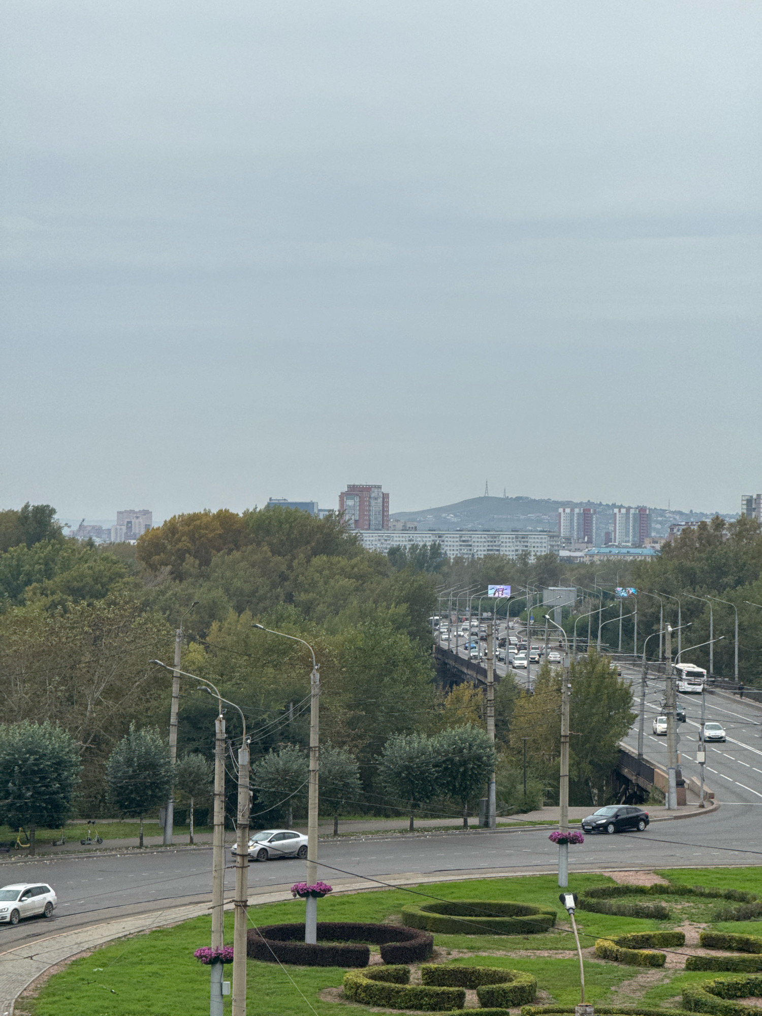 The image shows an overcast day in Krasnoyarsk, Russia, with a view of a multi-lane elevated road or bridge carrying traffic, bordered by trees and the city skyline in the distance. The traffic appears moderate, with cars and a bus visible on the roadway. On the right side of the road, a white bus is traveling away from the viewer, and a white sedan is traveling toward the viewer. On the left side of the road, a dark sedan is turning. In the foreground, there is a green grassy area with decorative manicured bushes and lampposts. Several scooters are parked on a sidewalk in the middle ground. The background features a dense canopy of trees, with several multi-story buildings and a distant hill with a radio tower visible beyond the trees. The sky is a uniform, pale gray, suggesting cloudy weather.