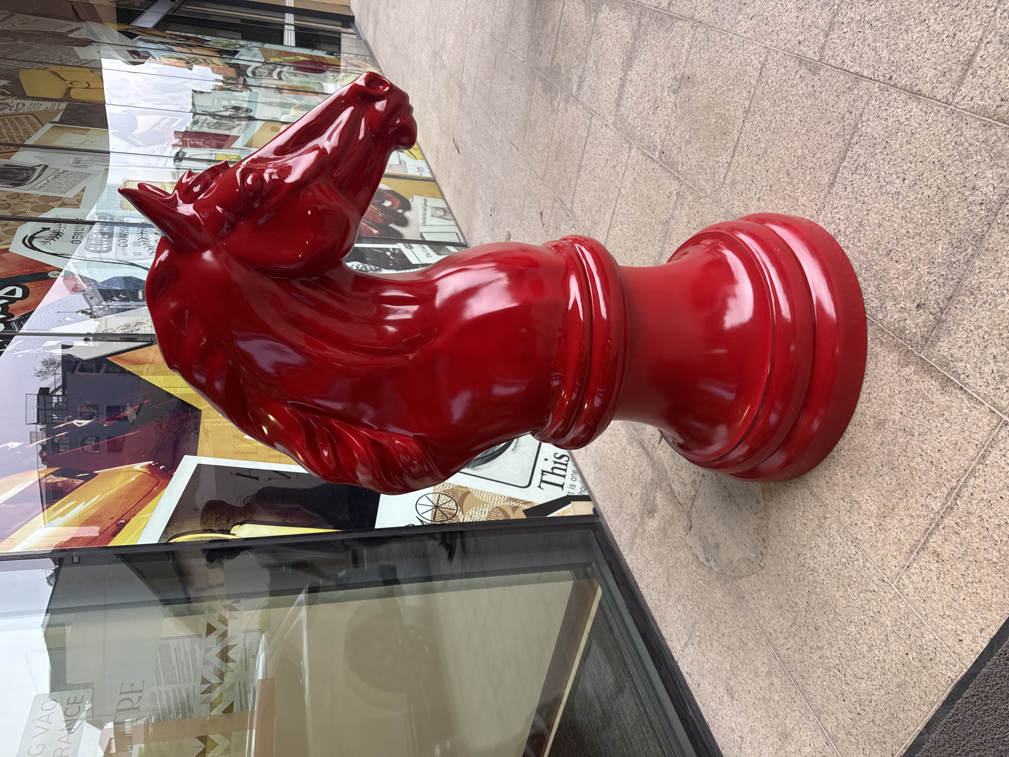 A glossy red, oversized chess knight statue stands on a tiled sidewalk next to a glass-fronted building with reflective posters in Hà Nội, Vietnam. The statue is the central focus. The building behind the statue appears to be a store or gallery, judging from the posters visible through the glass. The background consists of other buildings.