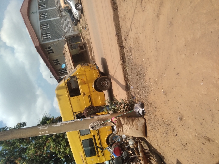 This is a daytime street scene in Agege, Nigeria, under a partly cloudy sky.
In the left foreground, a red and black motorcycle is partially visible, parked on a reddish-brown dirt and gravel surface. Adjacent to it stands a light-colored utility pole with white markings. Leaning against shrubbery near the pole is a large, light-brown sack.
A prominent yellow commercial van, a type of minibus, is positioned on a paved road that traverses the mid-ground from left to right. The van faces right and is slightly angled away from the viewer. A male individual, wearing a patterned shirt and blue trousers, stands next to the driver's side of the yellow van, facing away from the camera. The paved road features a raised curb separating it from the unpaved shoulder.
In the background, a multi-story building with a reddish-brown tiled roof and a grey facade dominates the right side. It features multiple white-framed windows and a distinctive white crest or logo near the roofline. A blue satellite dish is mounted on a pole near this building. Several parked vehicles, including a dark sedan and a silver sedan, are visible on the road or adjacent parking area near the main building. A smaller, darker-roofed structure is also visible behind the yellow van.
The immediate ground in the foreground and the road shoulder consists of reddish-brown dirt, gravel, and scattered debris, transitioning to a paved road surface.