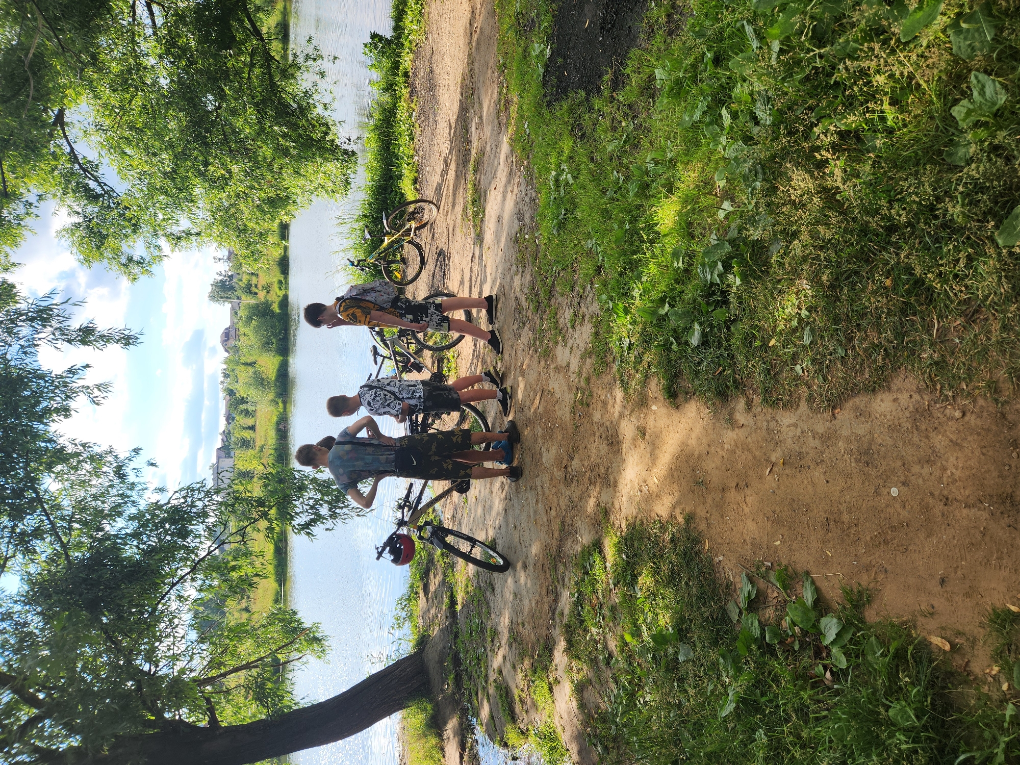 Three young males, appearing to be adolescents, stand near three bicycles by the edge of a tranquil body of water, possibly a lake or river, in Vniissok, Russia. The scene is set under the dappled shade of large, leafy trees. The ground is a mixture of sand and dirt, with patches of green grass and vegetation. In the background, across the water, a suburban landscape with houses and trees is visible. The sky is bright with scattered clouds, indicating a sunny day. One of the males is wearing a t-shirt with a camouflage pattern and dark shorts, while another wears a patterned t-shirt and dark shorts. The third male is seen from behind, wearing a t-shirt and dark shorts. One of the bicycles has a red helmet resting on its handlebars. The overall atmosphere suggests a casual outdoor activity, likely a bike ride near a natural setting within or on the outskirts of Vniissok.