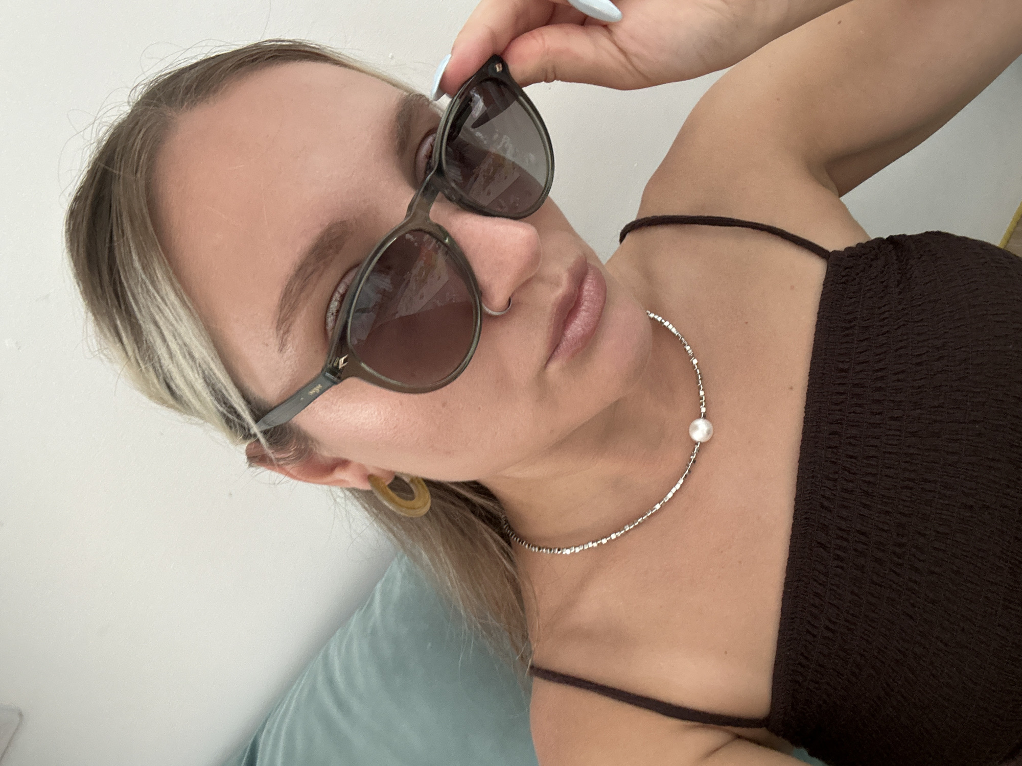 The image shows a close-up of a young woman from the chest up, wearing sunglasses, a brown smocked top, a silver necklace with a pearl, and a hoop earring. She is also wearing a nose ring. Her right hand is touching the arm of her sunglasses. The background is a light-colored wall and a section of a light blue cushion. There are no indications of the location being Vsevolozhsk, Russia.