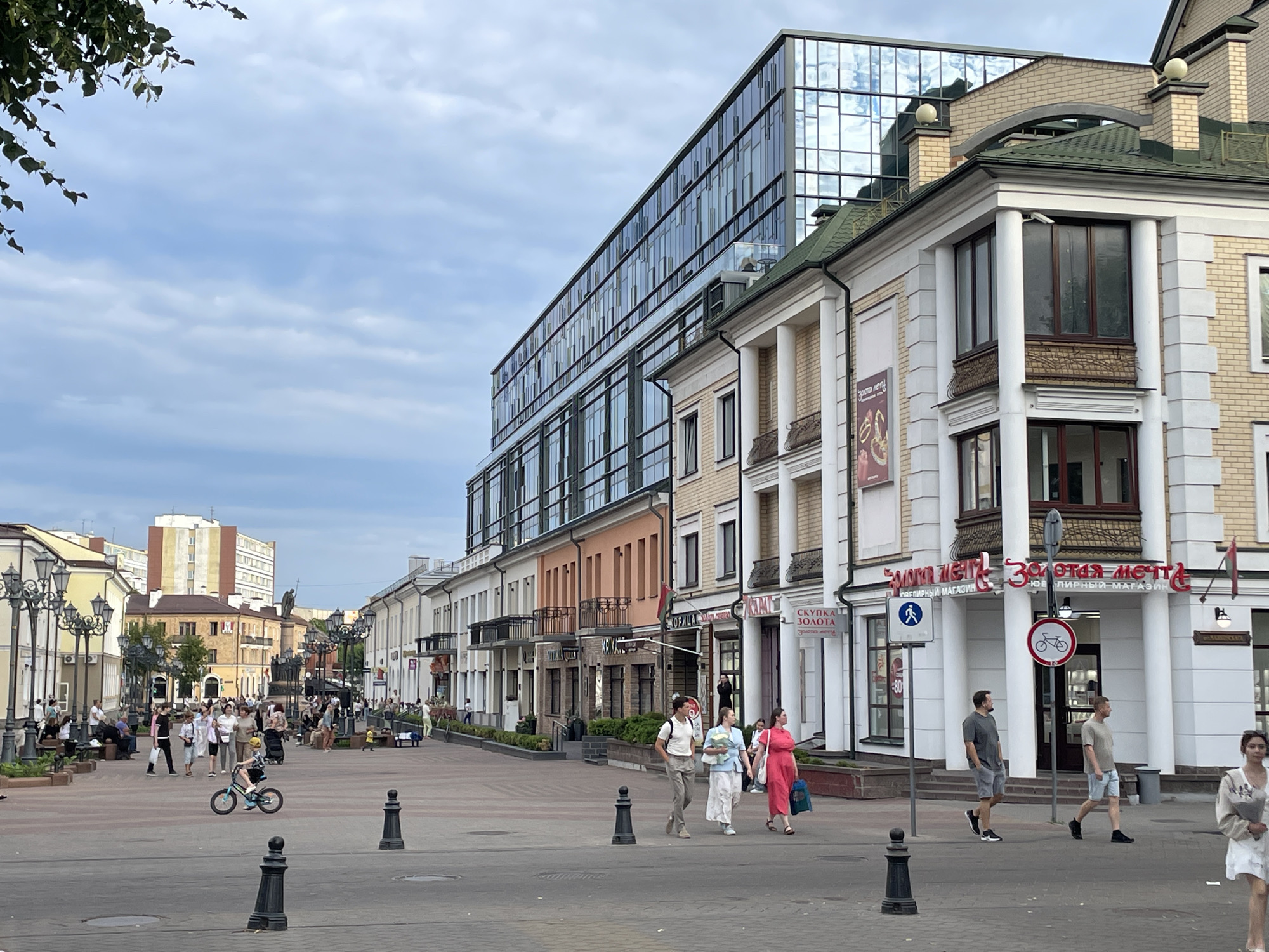 A medium-angle shot captures a street scene in Brest, Belarus, on a cloudy day. The bustling street is lined with shops and buildings, some with traditional architecture and others featuring modern glass facades. In the foreground, a young boy rides a bicycle, while several adults walk and socialize on the paved pedestrian area. The buildings display various architectural styles, including classical elements with columns and balconies, as well as a contemporary building with a large glass exterior reflecting the sky. Benches and lampposts are scattered along the sidewalk, suggesting a public square or promenade.
The activity appears to be a typical day in a city center, with people enjoying a stroll and shopping. The time of day seems to be late afternoon or early evening, given the soft light and the people's attire. The weather is overcast, but not raining. Visible text on the buildings includes "ЗОЛОТАЯ МЕЧТА" (Golden Dream) and "СКУПКА ЗОЛОТА" (Gold Purchase), indicating jewelry or pawn shops. The overall atmosphere is one of everyday urban life, with a mix of leisurely activity and commercial engagement.