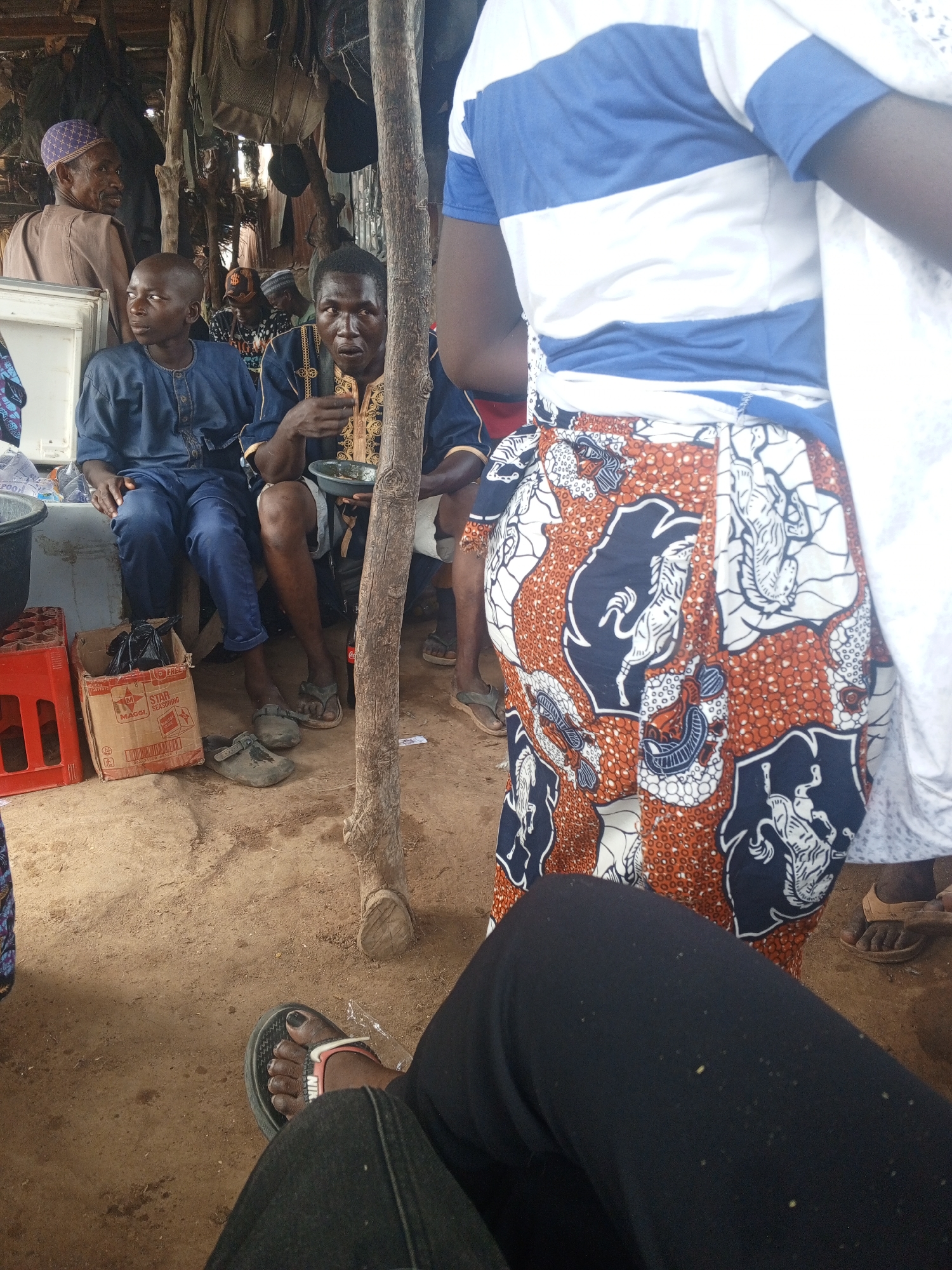 A medium shot captures a group of individuals in what appears to be an outdoor setting, possibly a gathering or public space in the city of Unknown, Unknown. A young man in a blue traditional outfit and an older man holding a bowl are sitting on a concrete block. Next to them, an older man is seated. Other individuals are visible in the background, some seated and some standing. A tree trunk stands prominently in the foreground, partially obscuring the view. In the foreground, a person is wearing black pants and a Nike sandal. A woman wearing a striped shirt and patterned skirt appears to be in the shot. Various containers, boxes, and personal items are scattered on the ground, suggesting a casual and relaxed atmosphere. The ground is bare earth, and the overall setting appears informal and unadorned.