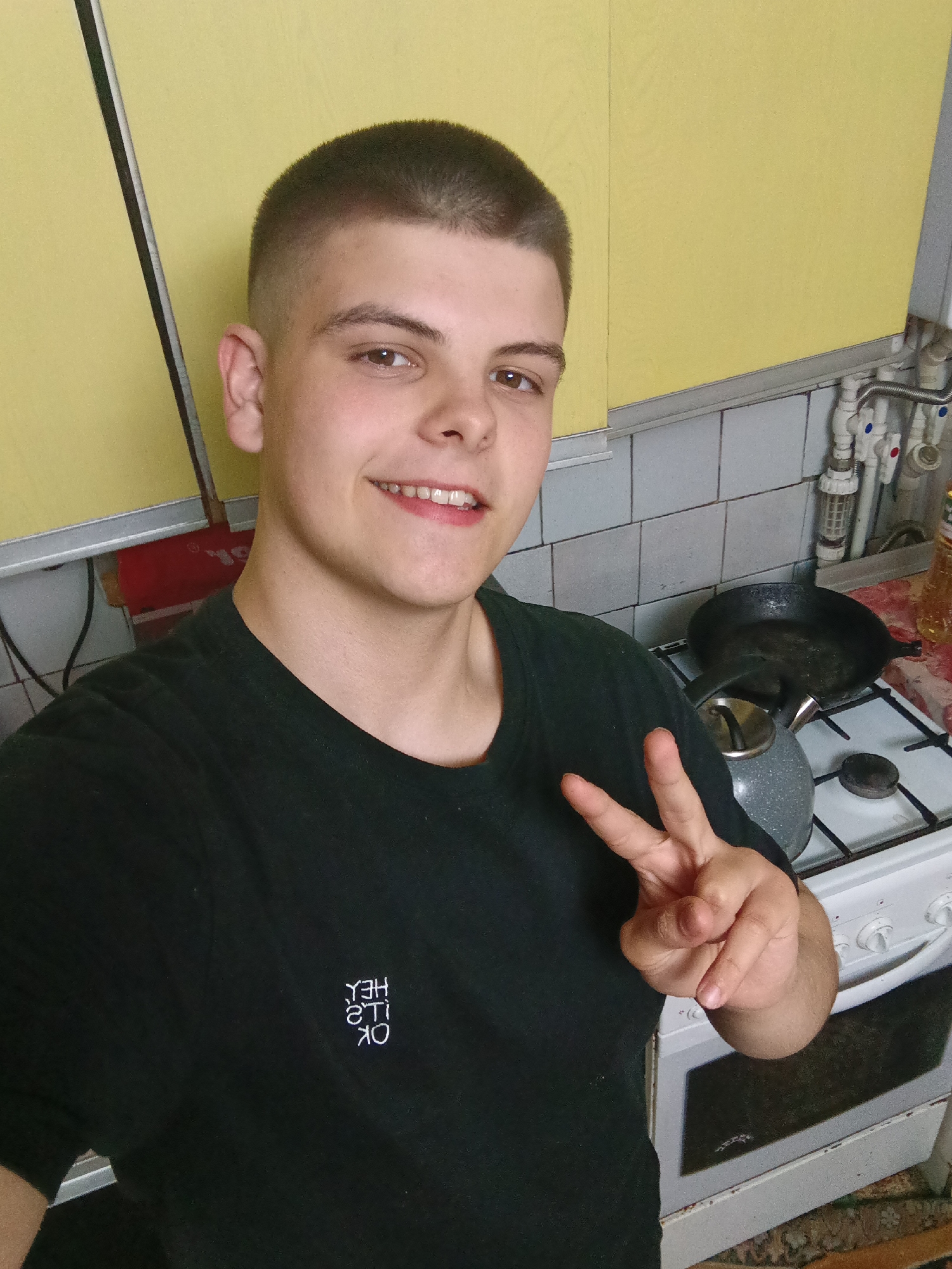 A young man takes a selfie indoors, likely in a kitchen. He has short, dark hair and is wearing a dark t-shirt with white writing on the chest. He smiles and gives a peace sign with his right hand. Behind him is a kitchen area with yellow cabinets and tiled walls. A stovetop with a kettle and a frying pan is visible. A bottle of oil sits on the counter. No information identifies the scene in the city of Красный Маяк, Russia.