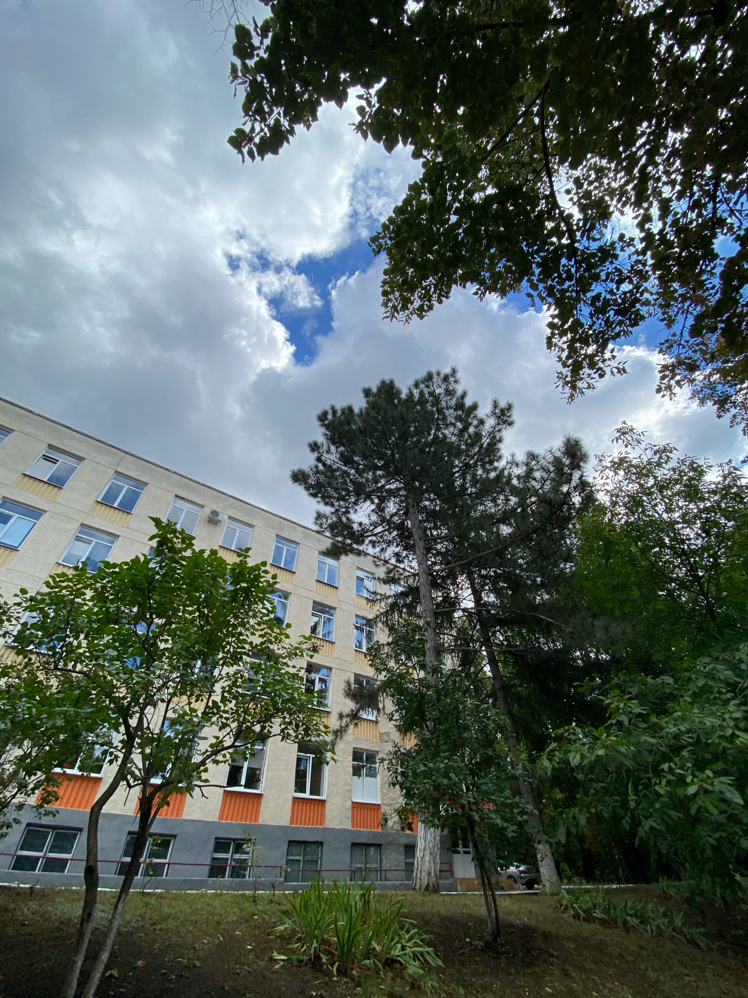 The image shows a low-angle view of a building with multiple floors, featuring a pale yellow facade and orange and grey accents. The building has numerous windows, some open, and an air conditioning unit visible on one of the upper floors. In the foreground and surrounding the building are various trees with green foliage, some casting shadows. The sky above is partly cloudy with patches of blue sky visible. The scene appears to be an exterior shot of a residential or institutional building.