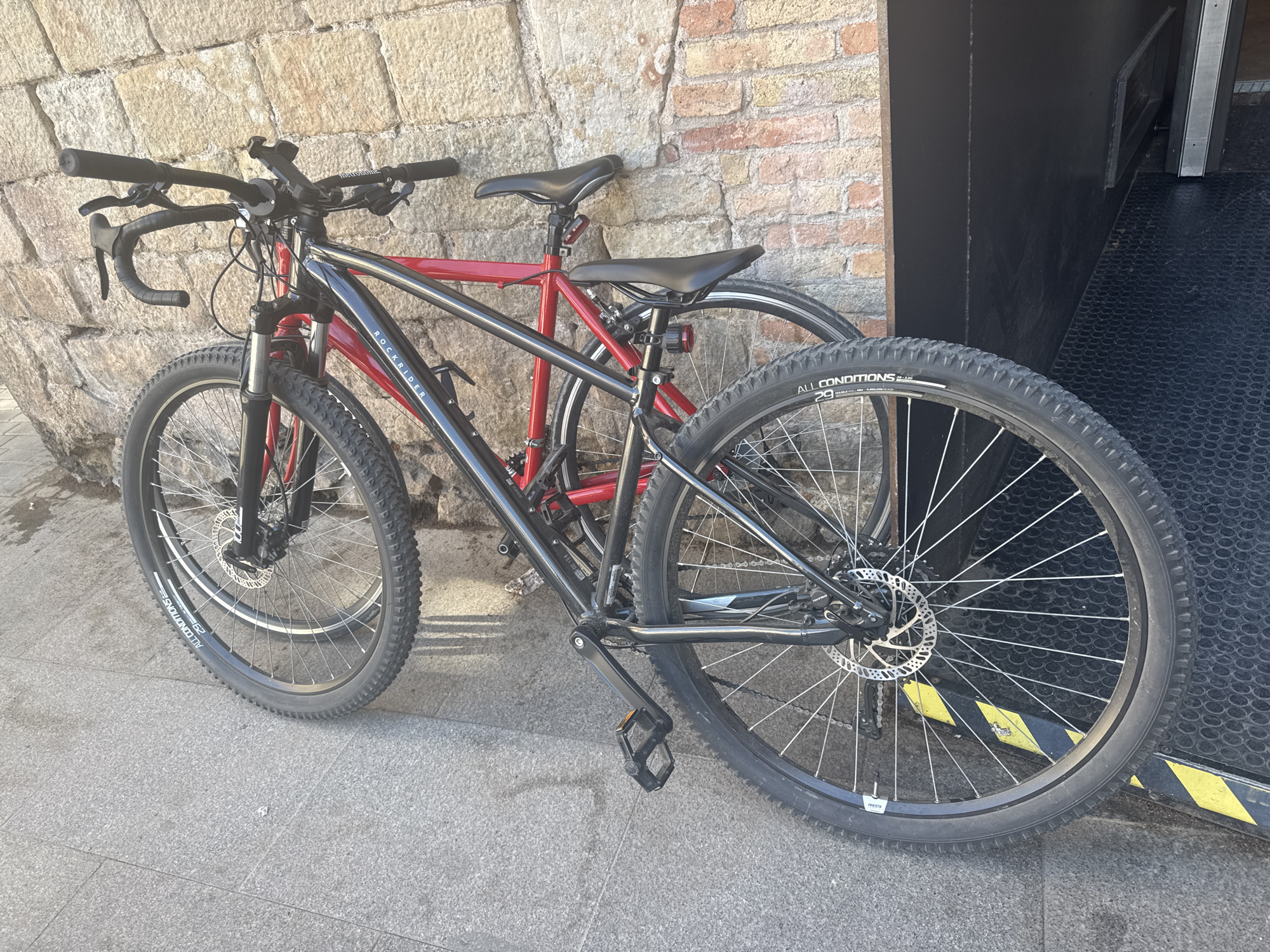 Two bicycles are parked side-by-side, leaning against an old, sun-drenched stone and brick wall. The setting appears to be a street or alleyway in Barcelona, Spain, given the provided context.
In the foreground, a black and red hybrid bicycle dominates the frame. Its frame features "ROCKRIDER" branding on the downtube, and a water bottle cage mount is visible. The handlebar setup is notable, featuring both flat bars with black grips and what appear to be drop bars, suggesting a customized configuration. A "metronic" branded phone mount is attached to the flat handlebar. The bike is equipped with knobby "ALL CONDITIONS" tires, marked as "29" inches and "S-2.20", with additional text indicating "DOUBLE WALL" and "TUBELESS READY." Both front and rear wheels have disc brakes, and a "PRESTA" valve is visible on the rear wheel. Standard platform pedals are attached, and a black saddle has a red rear light mounted beneath it.
Behind the front bicycle, a second, predominantly red bicycle is partially visible, also featuring a black saddle with a red rear light. The bikes rest on a grey concrete pavement. To the right, a dark, possibly metallic or rubberized surface, resembling a loading ramp or entrance, is visible, marked with distinctive yellow and black striped hazard tape along its edge. The scene is illuminated by bright daylight, casting clear shadows and indicating sunny weather.