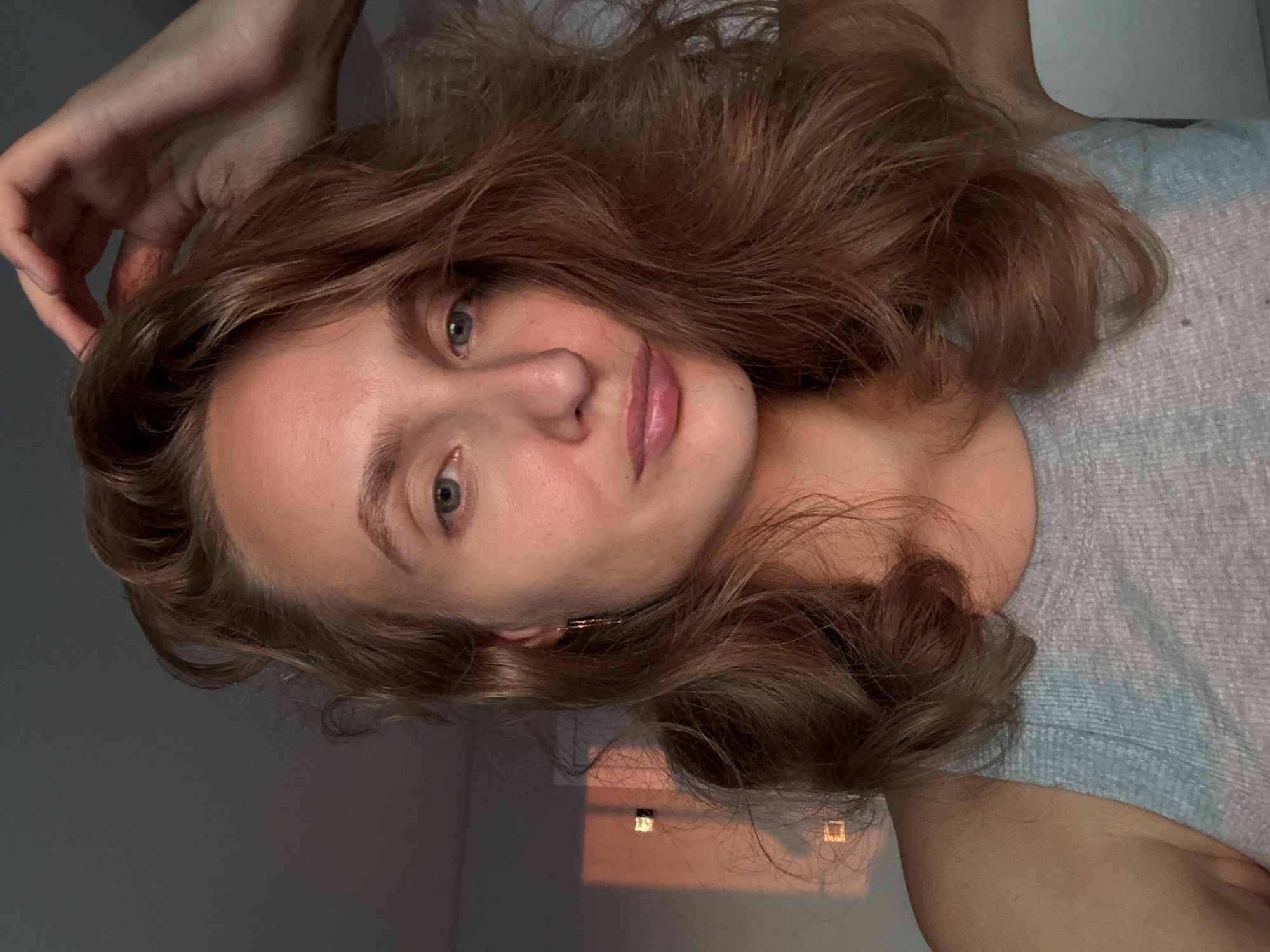 A close-up, selfie-style portrait of a woman with shoulder-length, wavy, auburn hair. She is looking directly at the camera with a neutral expression. Her right hand is raised and touching her hair. The woman is wearing a gray tank top. The lighting suggests it is either sunrise or sunset, casting a warm glow on her face and the wall behind her, which has geometric shadows from what appears to be a window. The background is a plain, light-colored wall. There is no indication of the city of Подшивалово, Russia in the image.