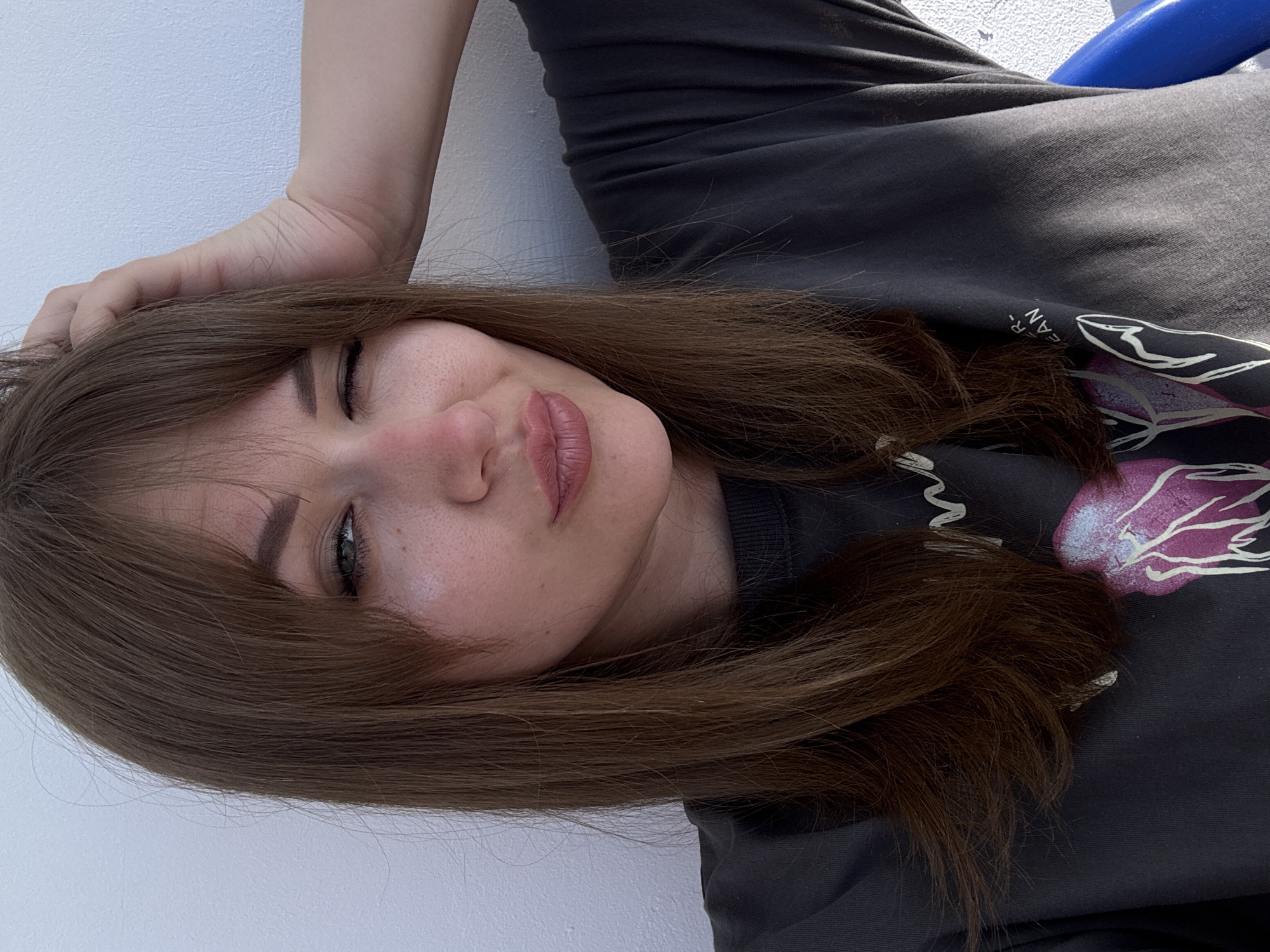 A young woman with light brown hair partially covering her face is lying down, looking directly at the camera with one eye closed in a playful wink. She has fair skin and pink lips. Her right hand is placed on her head, slightly lifting her hair. She is wearing a dark-colored t-shirt with a print on it. The background is a plain white surface. There is no evidence to indicate the location in Yevpatoriya, Ukraine.