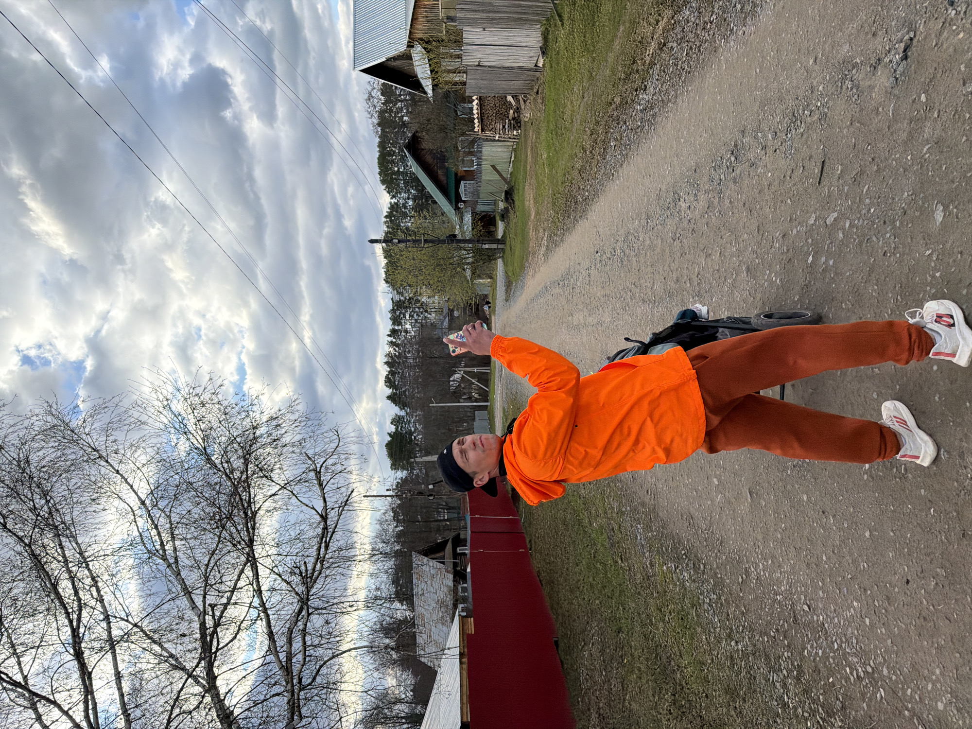 The image captures a male individual standing on a dirt road in a rural or semi-rural setting, consistent with Chaikovsky, Russia. He is dressed in a bright orange jacket, dark reddish-brown sweatpants, white sneakers with red stripes, and a dark beanie. He holds a smartphone in his right hand, pointing it slightly upwards and to his left, seemingly focused on something in that direction, perhaps taking a picture or video.
Behind his legs, a black bicycle wheel and frame are partially visible on the ground. The gravelly dirt road stretches into the mid-ground, flanked by a strip of green grass on the left. In the background, several rustic wooden buildings, possibly houses or barns, with corrugated metal roofs are visible, along with stacks of firewood. The landscape is dotted with trees, many still bare-branched, indicating early spring, while others show nascent green foliage. Overhead, power lines cut across a dramatic sky filled with a mix of light and dark grey clouds, with glimpses of blue sky peeking through. The time of day appears to be daytime, possibly late afternoon. No discernible text is present in the image, and the person's expression is neutral.