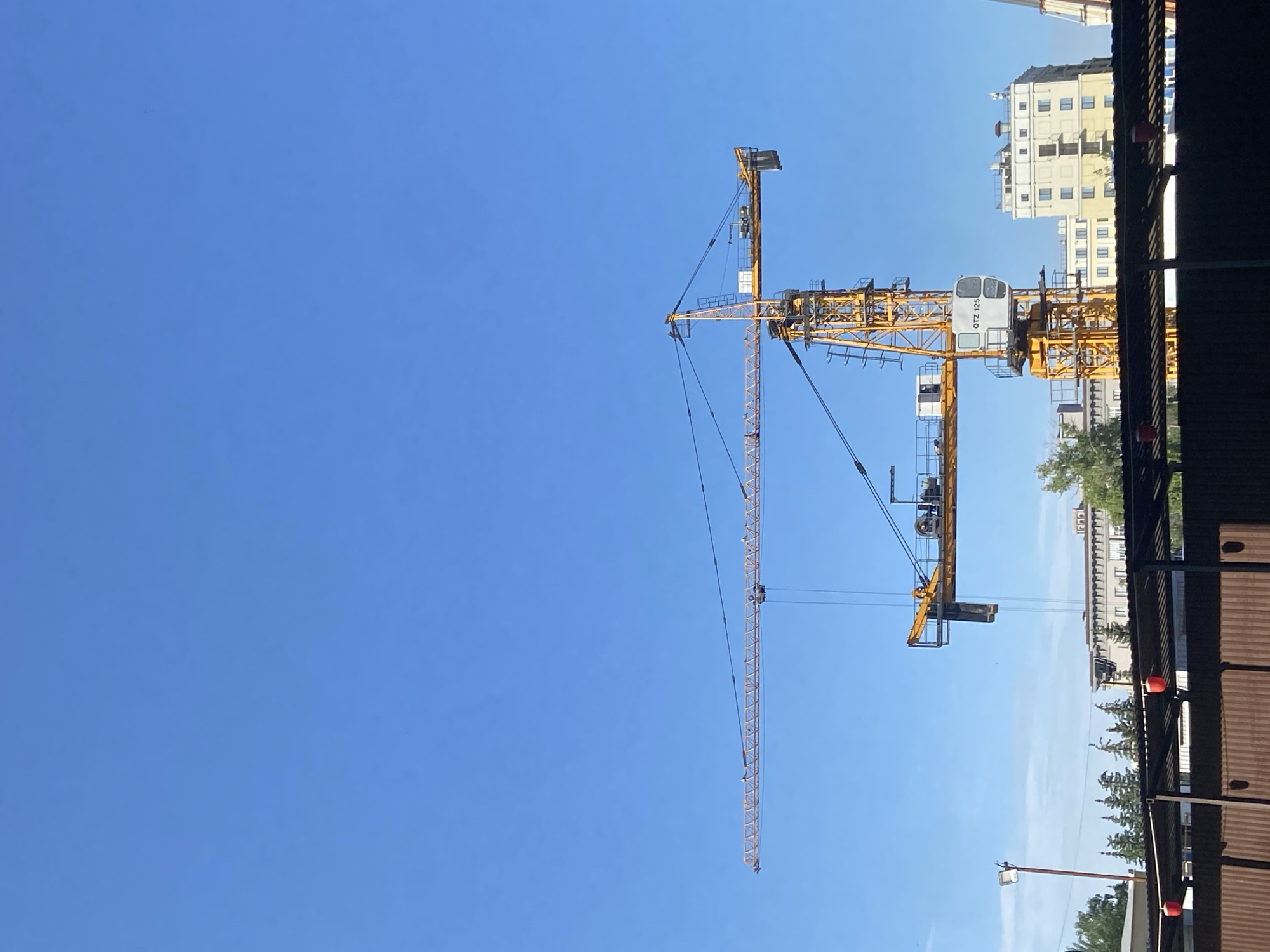 A yellow tower crane stands against a clear blue sky. The crane is located in Kazan, Russia. Part of the crane is obscured by the roof of a building. A tall building with a light exterior is visible near the crane's base. Trees and streetlights are also visible near the base of the crane.