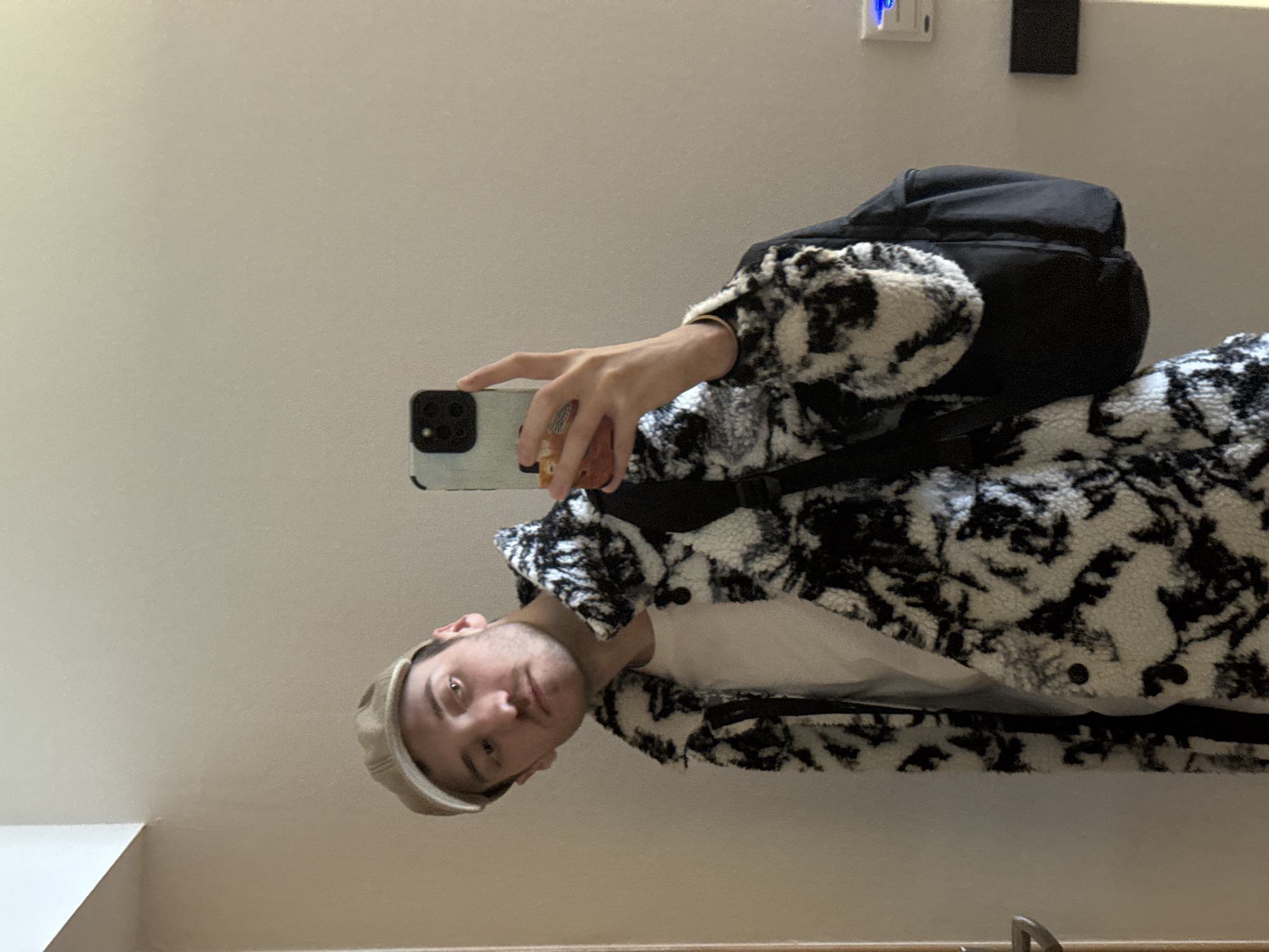 The image displays a young adult male taking a mirror selfie indoors in Kazan, Russia. The male, positioned slightly off-center and facing the camera with his head tilted, wears a light-colored baseball cap, a white t-shirt, and a black and white patterned sherpa-style jacket with visible black buttons. A dark-colored backpack is worn over his right shoulder. His right hand holds a smartphone, equipped with a light blue/grey case and a prominent black camera module, angled towards the mirror. A gold-colored bracelet is visible on his left wrist. The background consists of a plain, light-colored wall. A silver door handle is partially visible on a light-colored door frame on the left. On the right, a wall-mounted device with a blue glowing indicator light and a lower, dark rectangular object are present. The upper left portion of the image shows the intersection of the wall and ceiling.