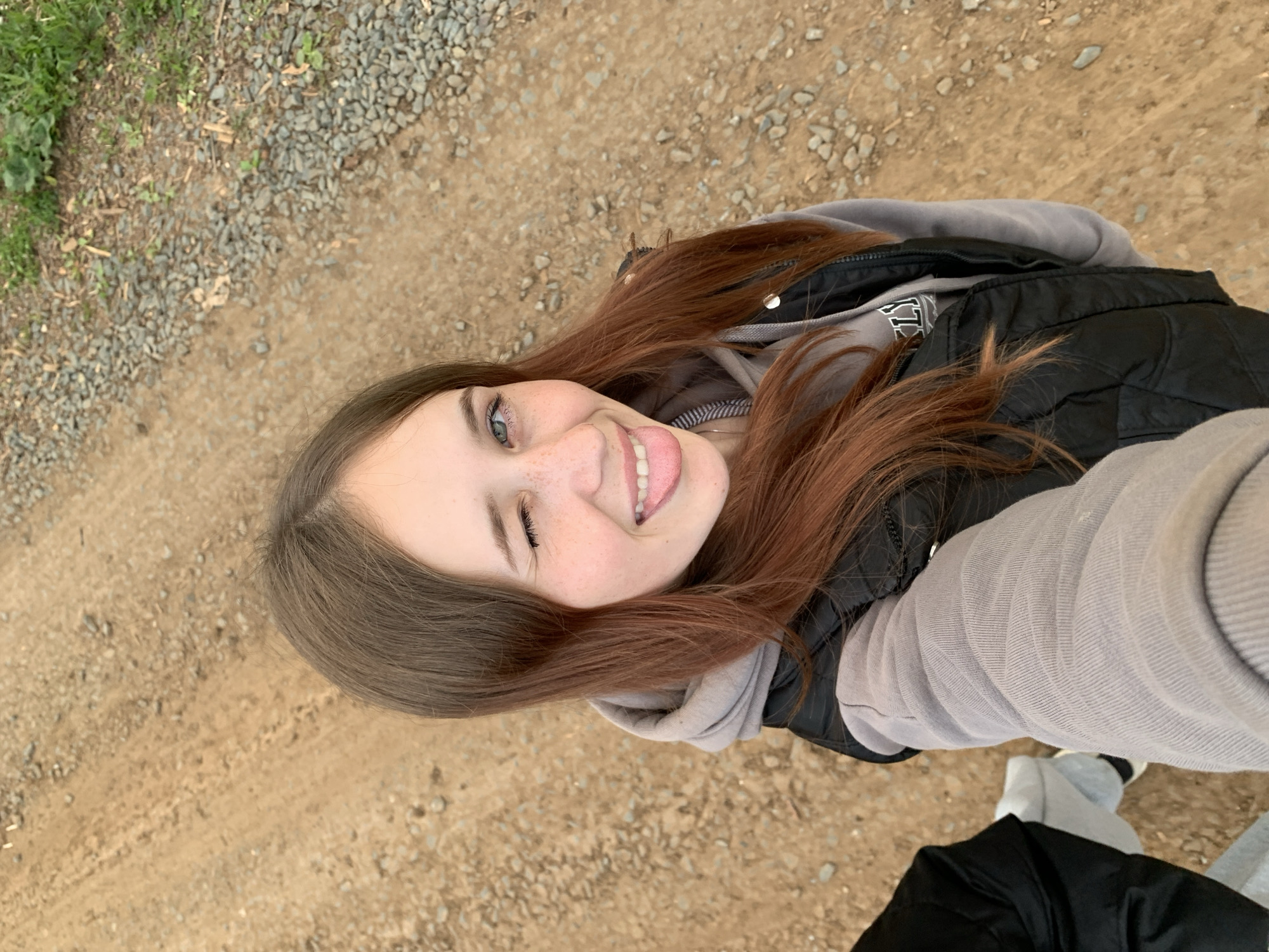 A close-up, high-angle selfie captures a young female outdoors in Фоки, Russia. The subject has long, reddish-brown hair, light-colored eyes, and visible freckles across her nose and cheeks. She is winking with her right eye (viewer's left), smiling, and extending her tongue. She wears a grey hooded sweatshirt, with partial letters "LS" visible on the chest area, layered under a black puffer vest. A silver ring earring is present in her left ear (viewer's right). The background consists of a textured dirt and gravel path, with small, grey stones interspersed with light brown soil. Sparse green vegetation is visible bordering the path on the right side of the frame. In the bottom-left corner, a partial view of another individual's black clothing and light-colored footwear is visible.