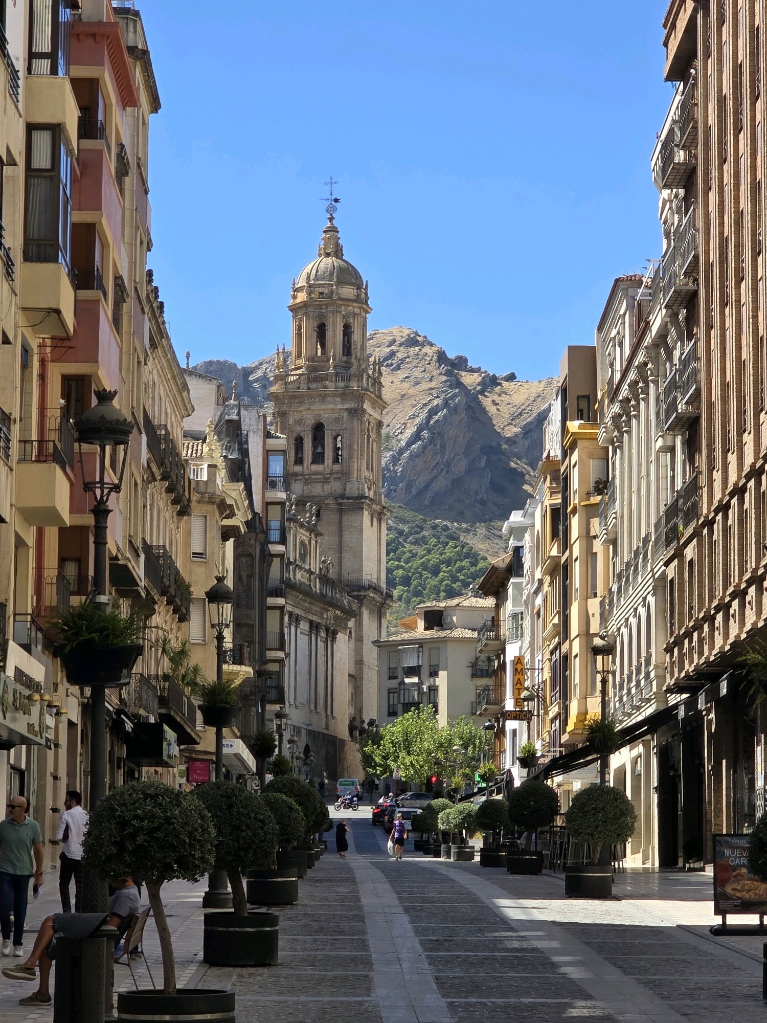 The image captures a street scene in Jaén, Spain, likely a pedestrianized area given the layout and presence of trees in planters. The perspective is from street level, looking down a wide avenue lined with buildings.
On the left, a building with colorful facades, featuring balconies and windows, dominates the foreground. A lamppost with decorative metalwork and a potted plant is situated prominently on this side. Further down the street, more buildings with balconies and awnings are visible.
On the right, a tall, narrow brick building with numerous windows and balconies stands out. This building appears to be a commercial establishment, with awnings extending over the ground floor.
In the mid-ground, a large, ornate bell tower with a dome and a cross on top rises above the surrounding buildings. This tower is a significant architectural landmark. Behind the tower and the buildings, a rugged, rocky mountain range with sparse vegetation forms the backdrop under a clear blue sky.
Several people are present in the scene. In the foreground on the left, a man in a green shirt and dark pants walks away from the camera, holding a document. Beside him, a man in a white shirt and dark pants stands, facing away. A man is seated on a chair in the shade of a tree planter. Further down the street, people are seen walking, and a woman in a dark dress is walking towards the camera. On the right side of the street, near the buildings, a person stands near a signboard. A motorcycle is visible in the distance on the right side of the street, indicating vehicle traffic further down the avenue.
The street itself is paved with cobblestones and lighter rectangular stones, forming a patterned walkway. Potted trees with rounded canopies are lined up along the pedestrian area, providing shade and decorative elements. Sunlight casts shadows across the pavement, indicating a bright, sunny day. The overall impression is a vibrant urban streetscape with a historical and architectural focus, set against a natural mountainous backdrop.