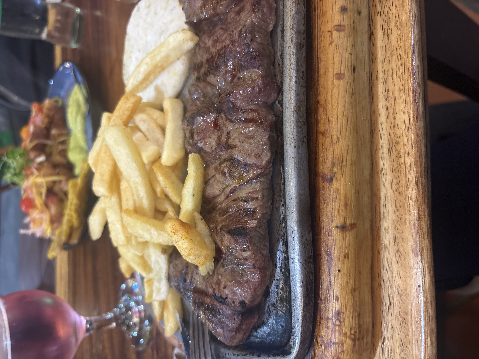 A close-up, tilted shot features a steak dish presented on a wooden table or surface. The steak, grilled with visible sear marks, is the central element, situated on a rectangular metal serving dish. Accompaniments include a mound of white rice and a pile of French fries. To the left, a small portion of what appears to be a salad or side dish is visible in a blue-rimmed bowl.
In the bottom left corner, part of a wine glass is present, suggesting a meal setting. The surrounding environment is dimly lit, adding a sense of intimacy or a restaurant setting.
While not directly visible in the image, based on the prompt, this scene is presumed to be located in the city of Guarne, Colombia.