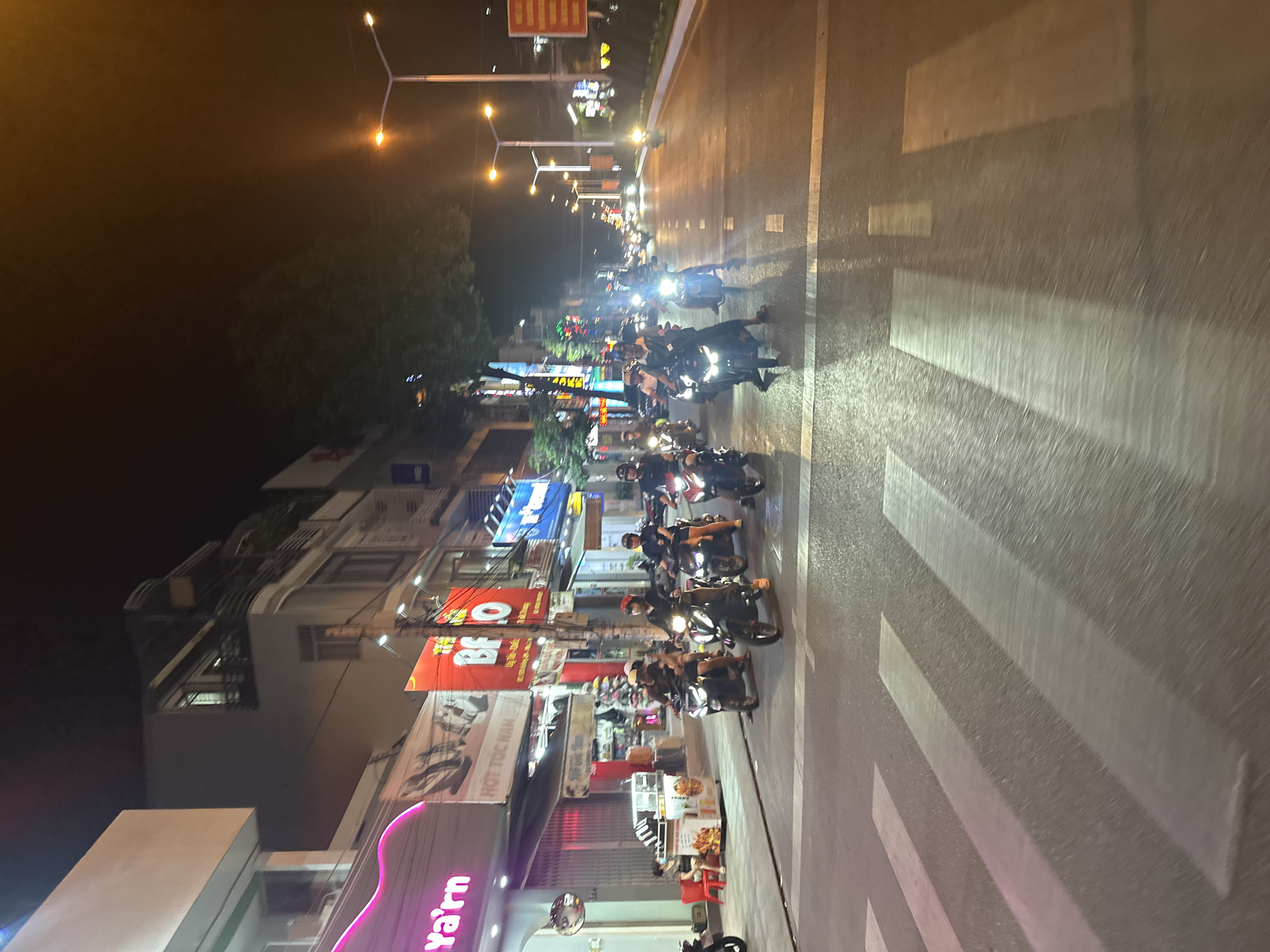 The image depicts a street scene at night in Bac Nha Trang Ward, Vietnam. A line of motorcycles is driving along the road, heading toward the camera. The buildings are multi-story with a variety of illuminated signs in Vietnamese. There are crosswalk markings on the road. Streetlights illuminate the scene.