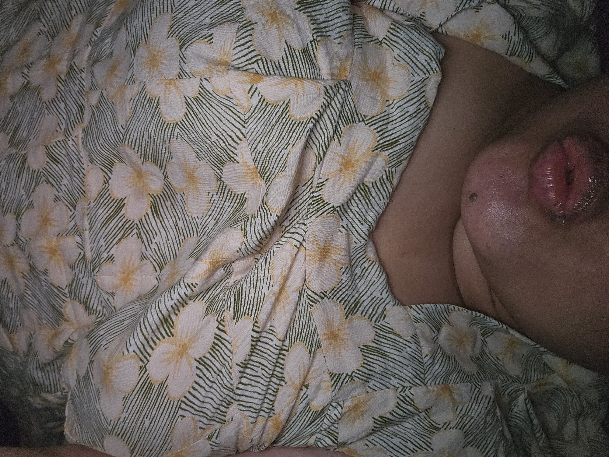 This media file is a close-up, overhead shot of a person's chest area, covered by a floral-patterned fabric. The fabric has a light background with scattered white flowers featuring yellow centers, and green, curved lines that suggest leaves or stems. The person's skin is visible on the right side of the frame, and a portion of their breast is also visible, with a mole present. The lighting is dim, suggesting an indoor setting, possibly a bedroom, at nighttime or in a dimly lit room. The person appears to be lying down, with the fabric draped over them. There is no visible text or specific location cues beyond the provided context of Lamongan, Indonesia. The image focuses on the texture and pattern of the fabric and the skin, creating an intimate and somewhat abstract composition.