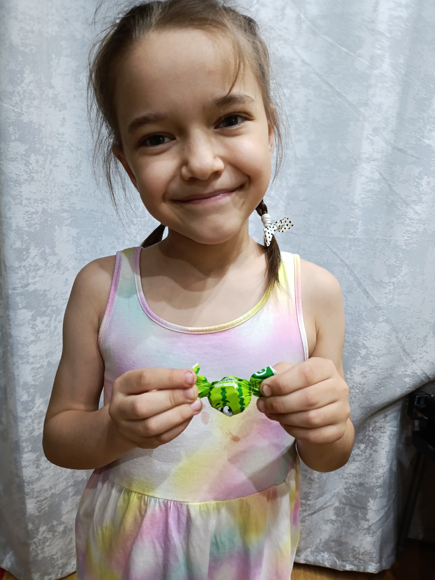 A young girl is holding a candy. She has fair skin, brown hair styled in two braids with polka dot bows, and is smiling. She is wearing a sleeveless dress with pastel tie-dye coloring. The candy she holds is wrapped in green paper with a watermelon design. The background features a light gray fabric backdrop and an out-of-focus dark object.
The location is Chaikovsky, Russia.