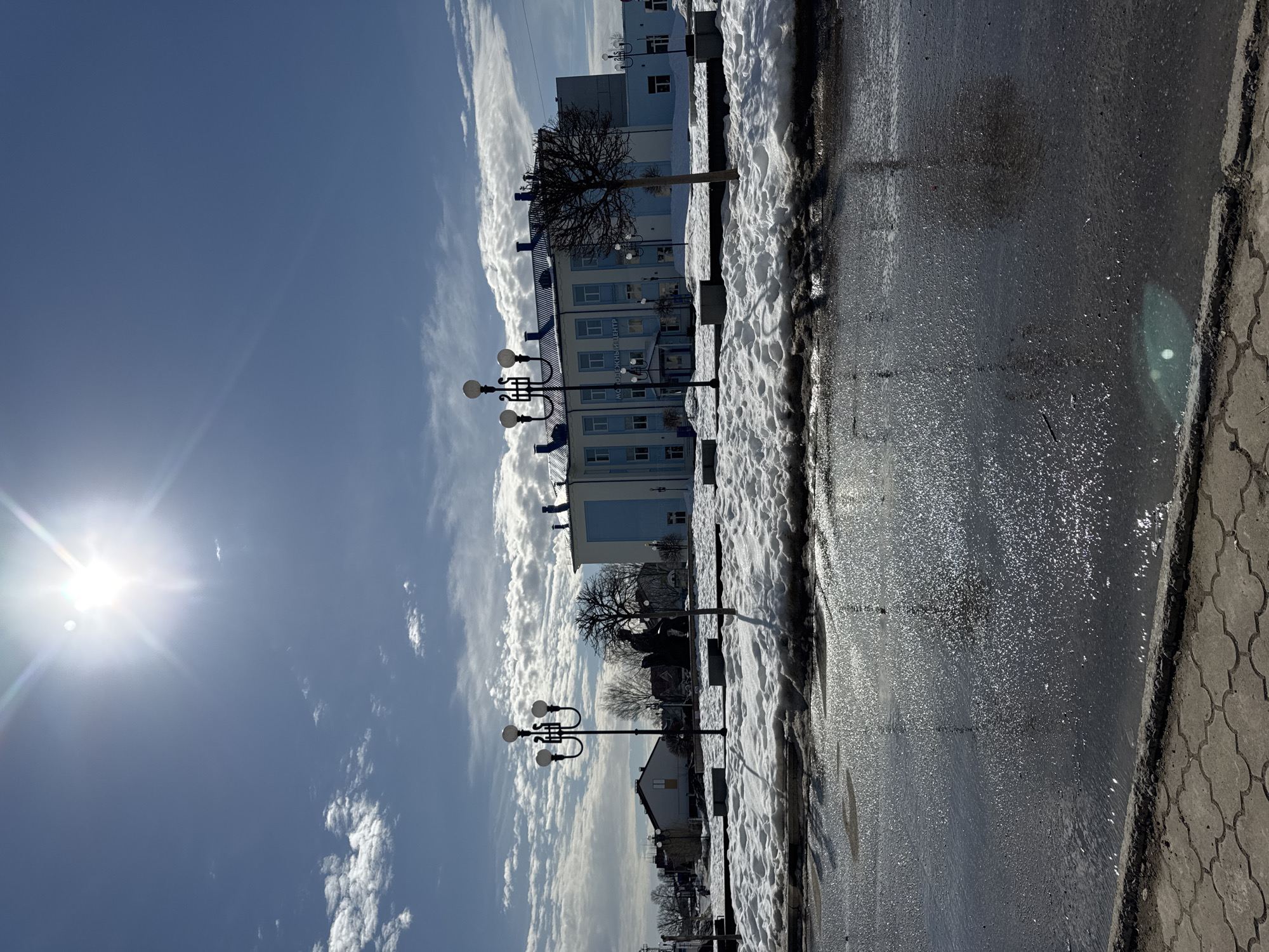 The image depicts an outdoor urban scene in the city of Chaikovsky, Russia, specifically focusing on the central square and the Youth Center building, during what appears to be late winter or early spring.
The foreground consists of a wet, dark paved surface with large puddles reflecting the bright overhead sunlight, indicating melting ice or recent precipitation. A grey paving stone curb is visible at the very bottom.
The midground features an irregular expanse of melting snow, exposing patches of dark ground. Within this area, two black lampposts, each with three spherical lights, are prominent. To the left, a dark, silhouetted statue of a seated figure, likely the monument to P.I. Tchaikovsky, is discernible. A small, bare deciduous tree stands to the right of the left lamppost. Several low concrete benches or planters are scattered throughout the snowy area.
In the background, the primary structure is a multi-story building identified by Russian text on its facade as "МОЛОДЕЖНЫЙ ЦЕНТР" (Youth Center). This building is painted light blue with white window frames and features a dark blue roof. Additional residential or commercial buildings are partially visible to the far left.
The sky is bright blue with scattered white cumulus clouds, more numerous towards the horizon and the lower left. The sun, a bright point of light with a prominent lens flare, is positioned in the upper left quadrant of the frame, illuminating the scene. No individuals or specific actions are observed within the frame.