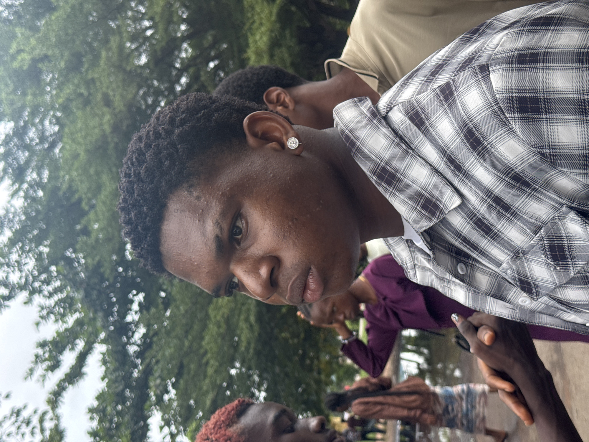 A close-up, profile view shows a young Black male in the foreground, facing left, with a blurred background of trees and other individuals. He has short, dark, curly hair and is wearing a checkered shirt with black and white patterns and a white undershirt. A silver earring with a circular design is visible in his left ear. His expression is neutral, with his lips slightly parted. His right hand is visible in the lower left of the frame, clasped with another hand, only partially visible and appearing to be that of another person. In the background, other individuals are seen, including a person with reddish-dyed hair to the left, and a person in a purple garment. The environment appears to be outdoors, possibly in a town or urban area, with lush green foliage suggesting a temperate climate or a heavily treed location within a city. The presence of puddles or wet ground in the mid-ground suggests recent rainfall. The overall scene is candid and appears to capture a moment between individuals in a public space within Ihiagwa, Nigeria.