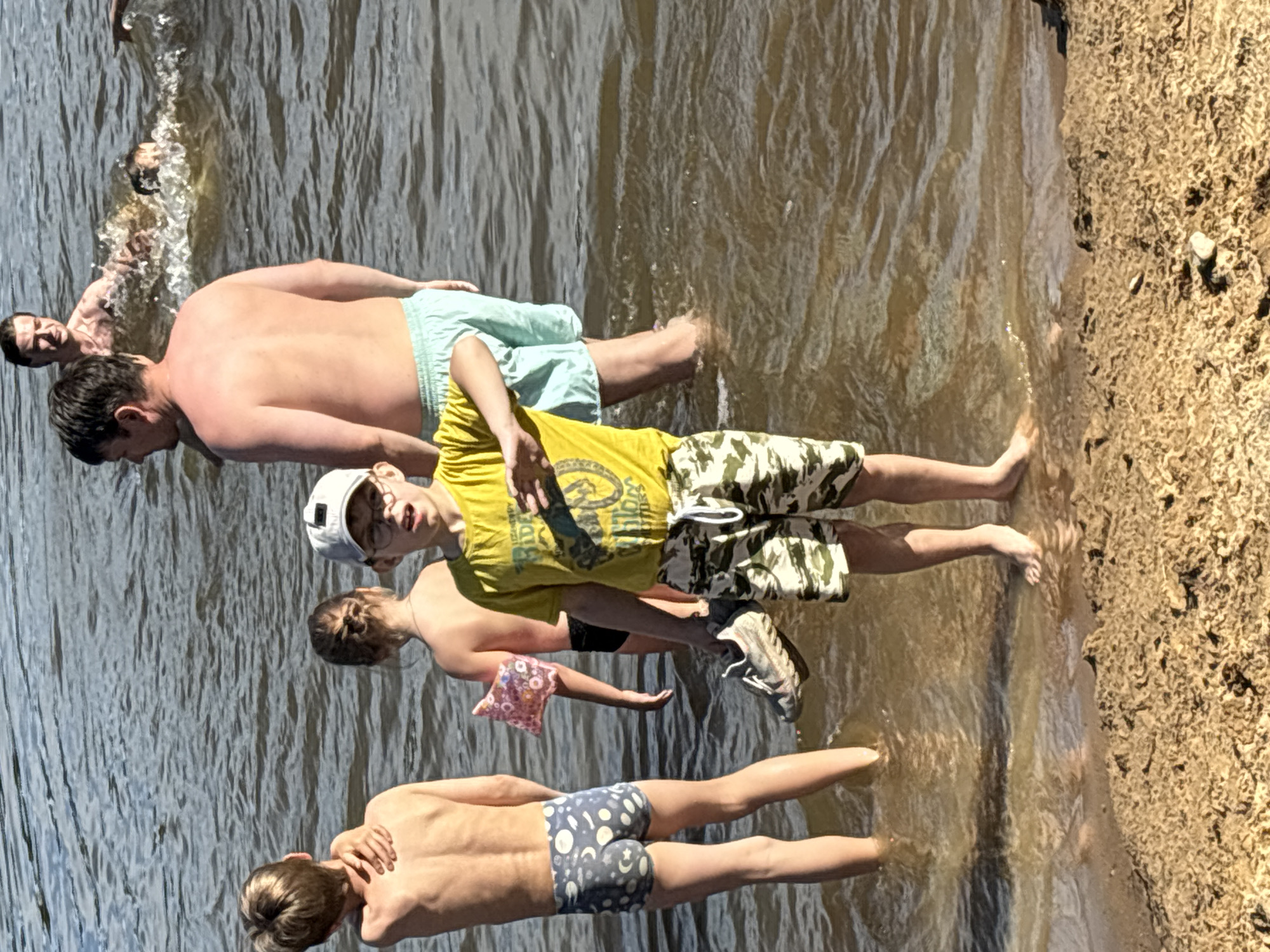 In Brilliant, Russia, a group of individuals are gathered at the edge of a body of water, likely a river or lake, on a sandy bank. The water is murky brown and appears calm with small ripples.
In the foreground, a young boy wearing a yellow t-shirt and camouflage shorts stands facing forward, holding a gray athletic shoe in his left hand. He has on a white baseball cap and glasses. To his left, another young boy in blue swim trunks with a moon and star pattern stands with his back to the camera. His left arm is resting on his shoulder. Behind him, a young girl with her hair in a bun is also facing away from the camera, wearing a dark swimsuit and a pink arm float with a floral pattern.
In the middle ground, a shirtless man with light-colored swim trunks stands facing the water. Further back, another shirtless man is partially submerged in the water, with another person swimming or playing in the water creating splashes. The background consists of more water and a sliver of land further away. The overall impression is a casual scene of people enjoying a warm day by the water.