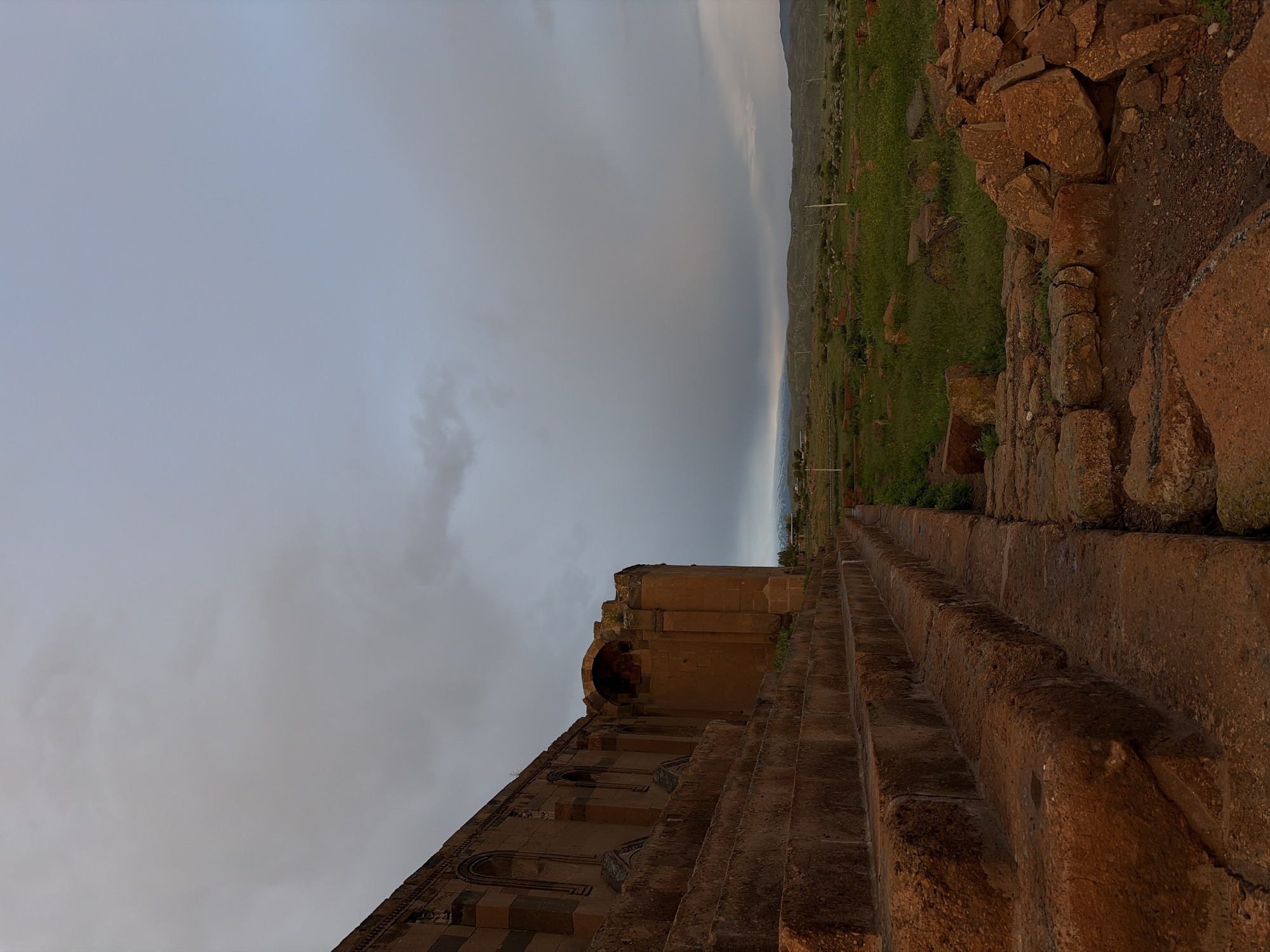 The image shows a partial view of a stone building, presumably a church or other religious structure, in Anipemza, Armenia. The structure has a curved archway, windows, and decorative patterns. Stone steps lead from the viewer towards the building. A landscape of green terrain and hills extends into the distance under a cloudy sky. A stone wall or path edges the green area.